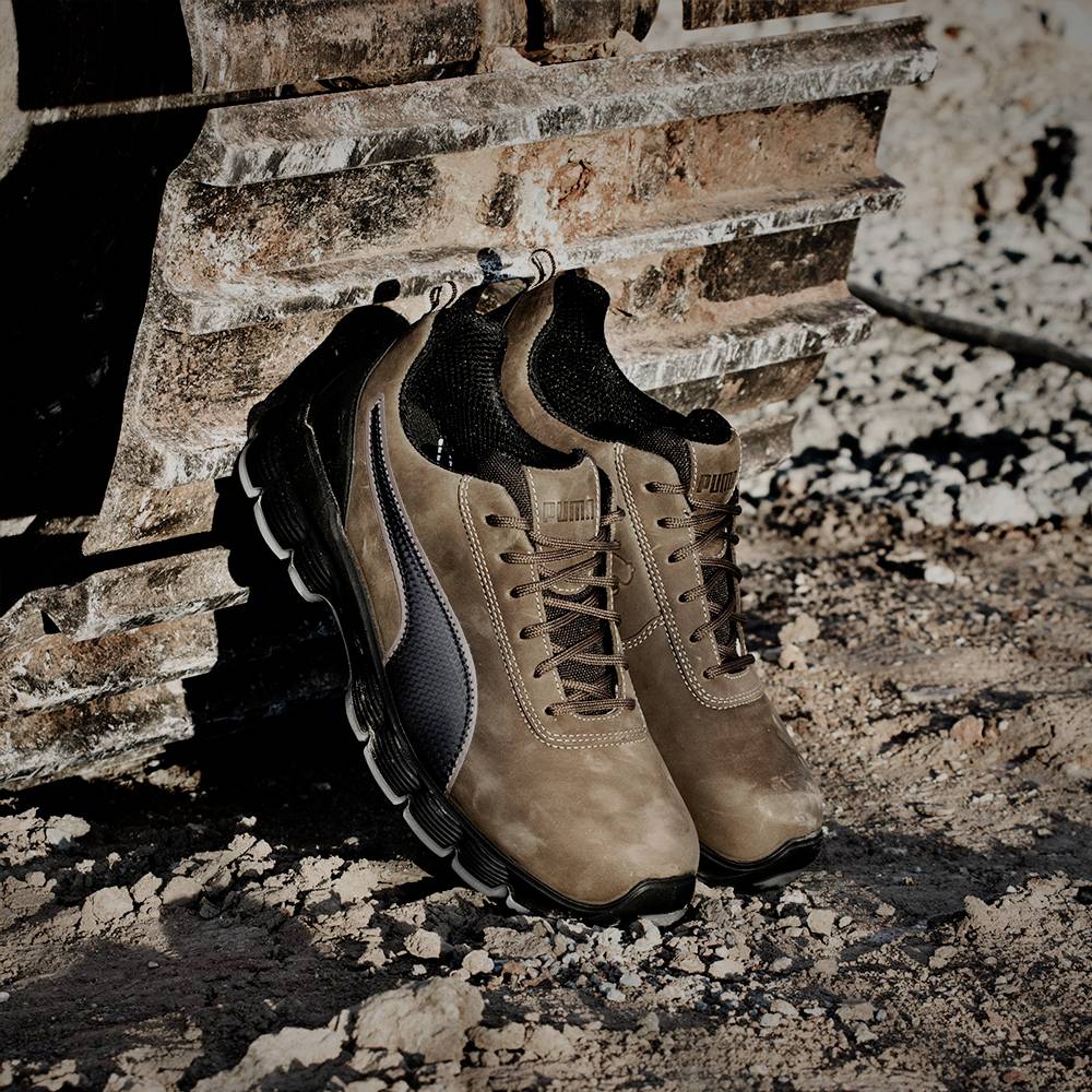 Deux chaussures de randonnée brunes avec semelle noire et lacets sont posées sur un sol rocailleux devant une construction métallique rouillée.