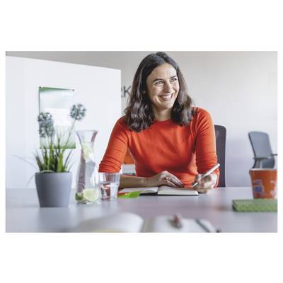 Femme souriante vêtue d'un pull rouge assise à un bureau, tenant un stylo et un carnet de notes dans une salle de réunion ou un bureau.
