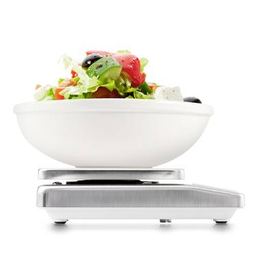 Un bol blanc rempli de salade, composé de tomates, olives, feta et de feuilles, est posé sur une balance de cuisine numérique.