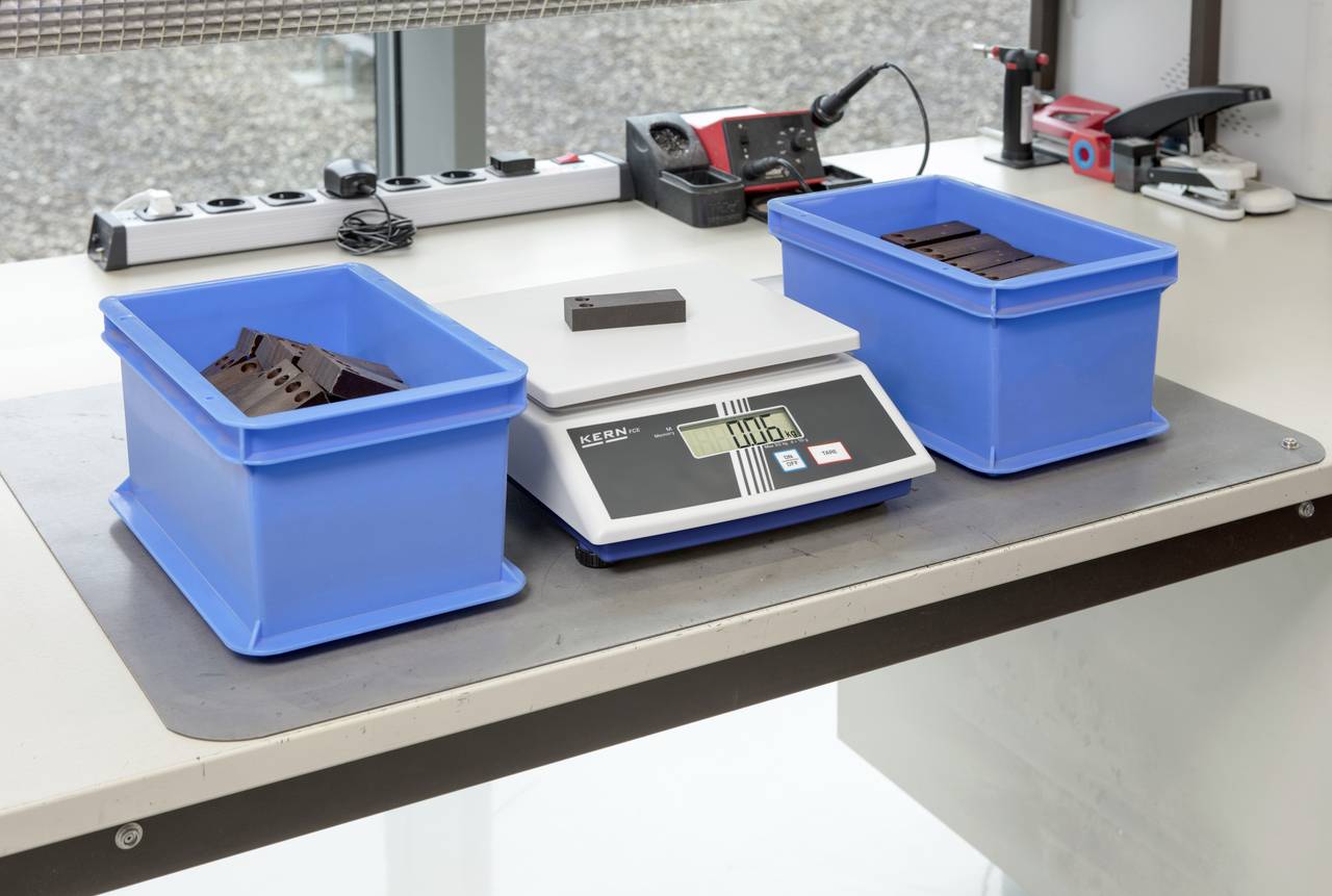 Deux boîtes en plastique bleu sur une balance. La balance indique '840,5 g'. En arrière-plan, différents appareils électroniques sont visibles.