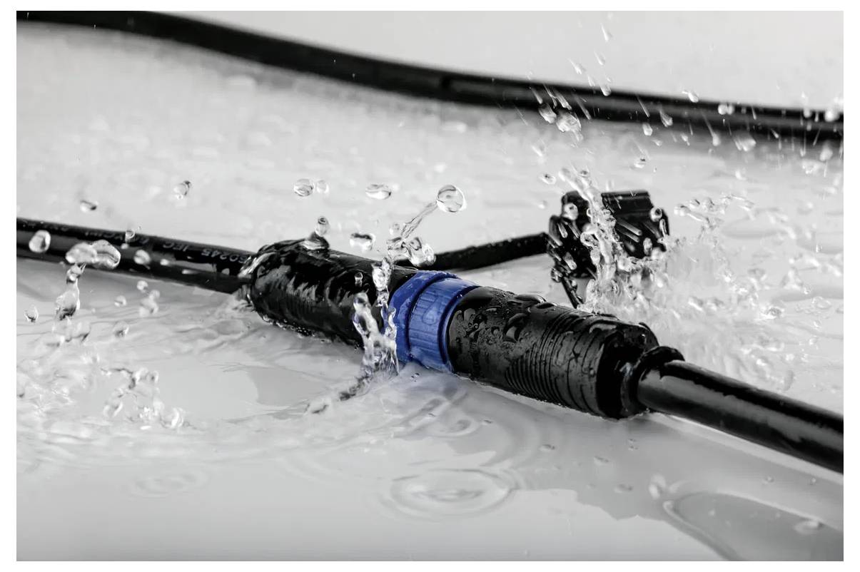 Un connecteur de câble noir marqué d'un anneau bleu est visible dans l'eau, démontrant sa caractéristique étanche. Des gouttelettes d'eau éclaboussent autour de lui.