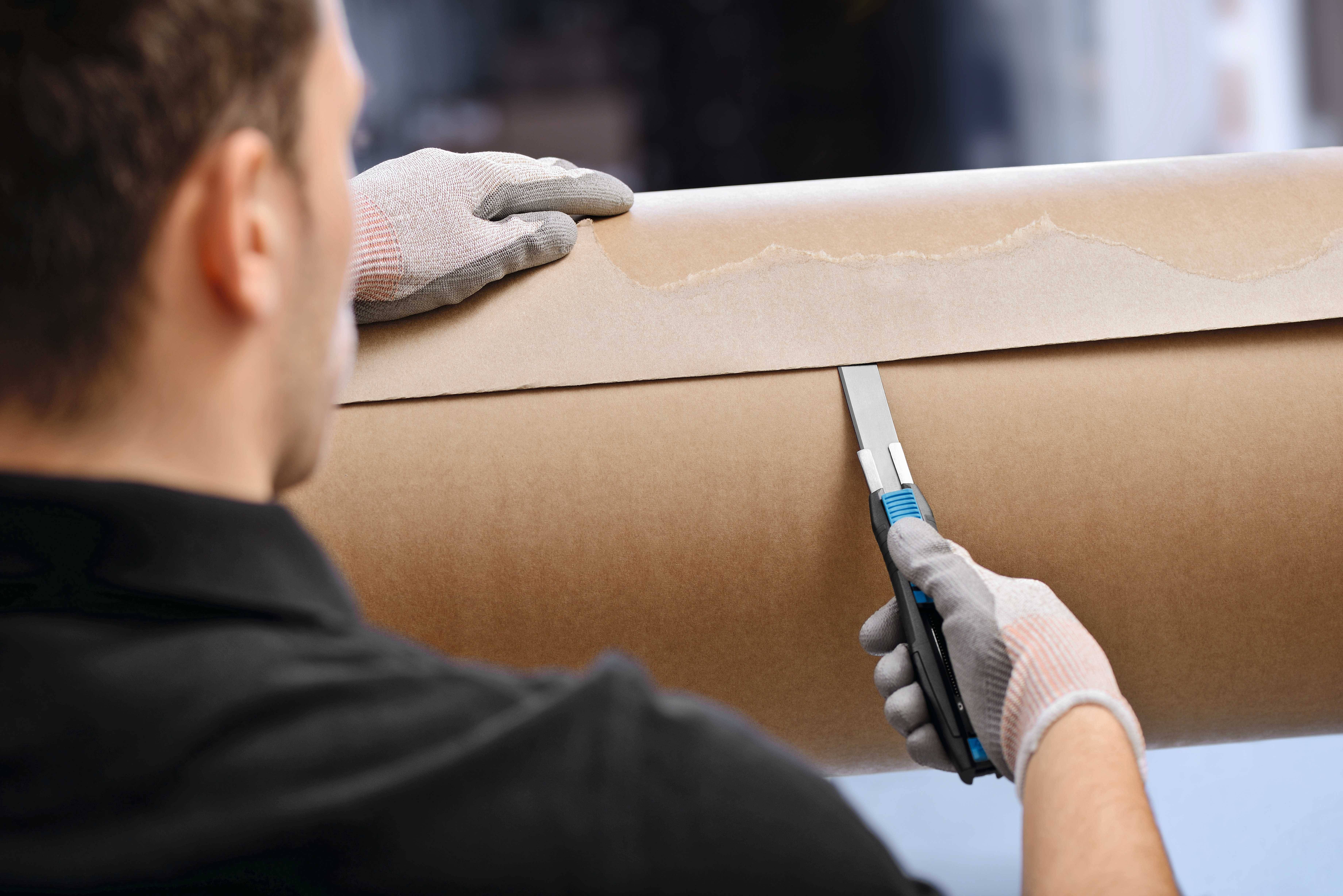 Une personne coupe une feuille de papier sur un grand rouleau à l'aide d'un cutter. Elle porte des gants de protection.