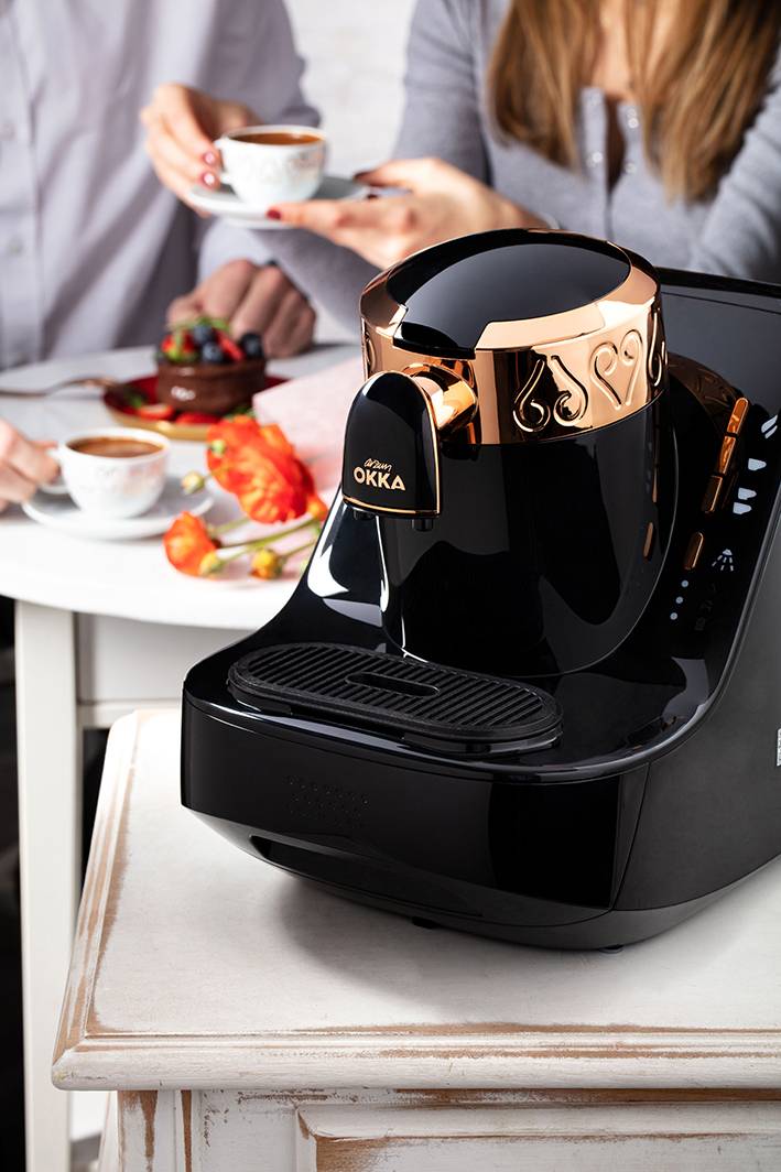 Machine à café noire sur une table, deux personnes buvant du café en arrière-plan.