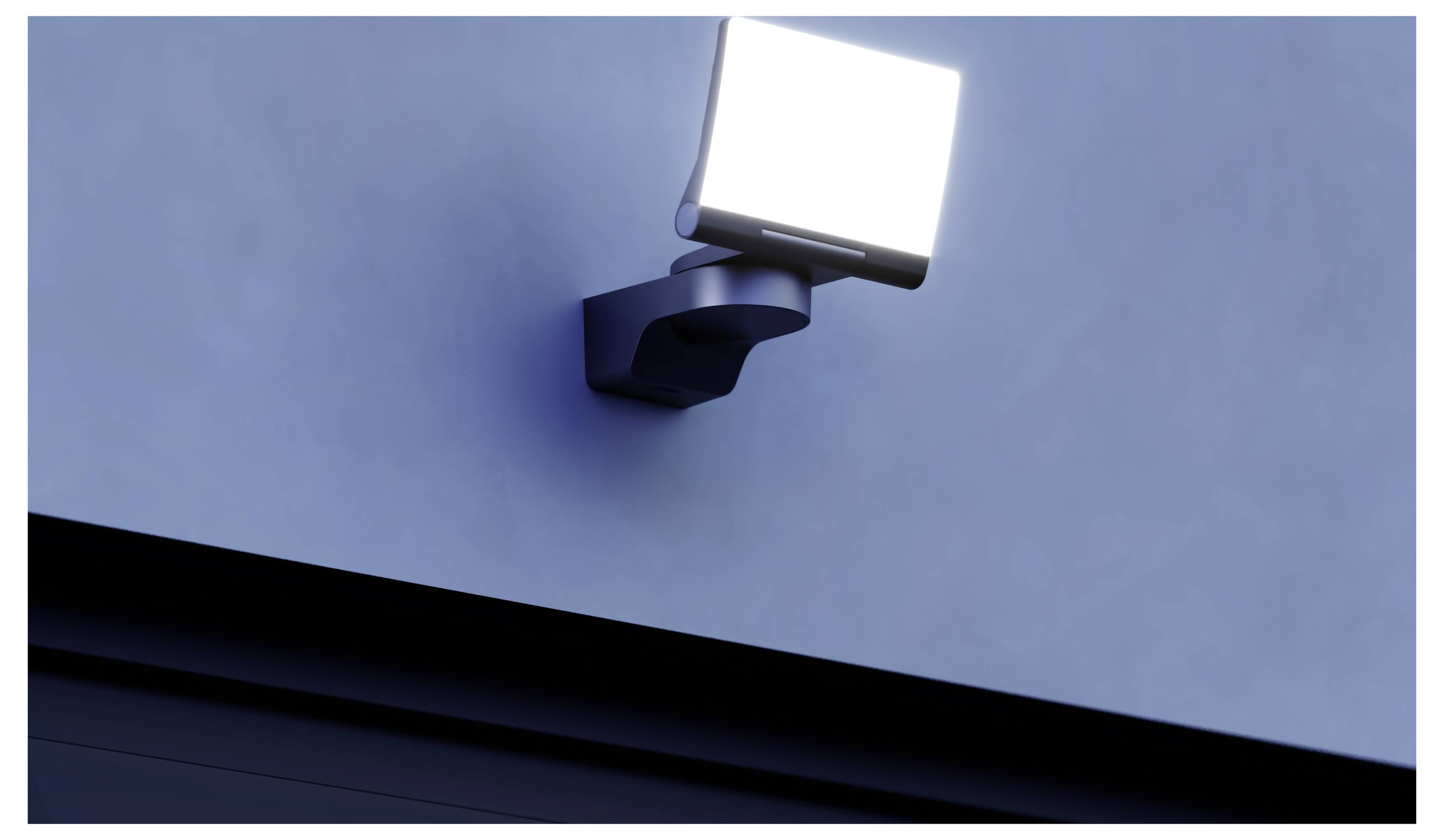 Luminaire de sécurité extérieur brillant monté en applique murale et illuminant un mur bleu au-dessus d'une ligne de toit sombre.