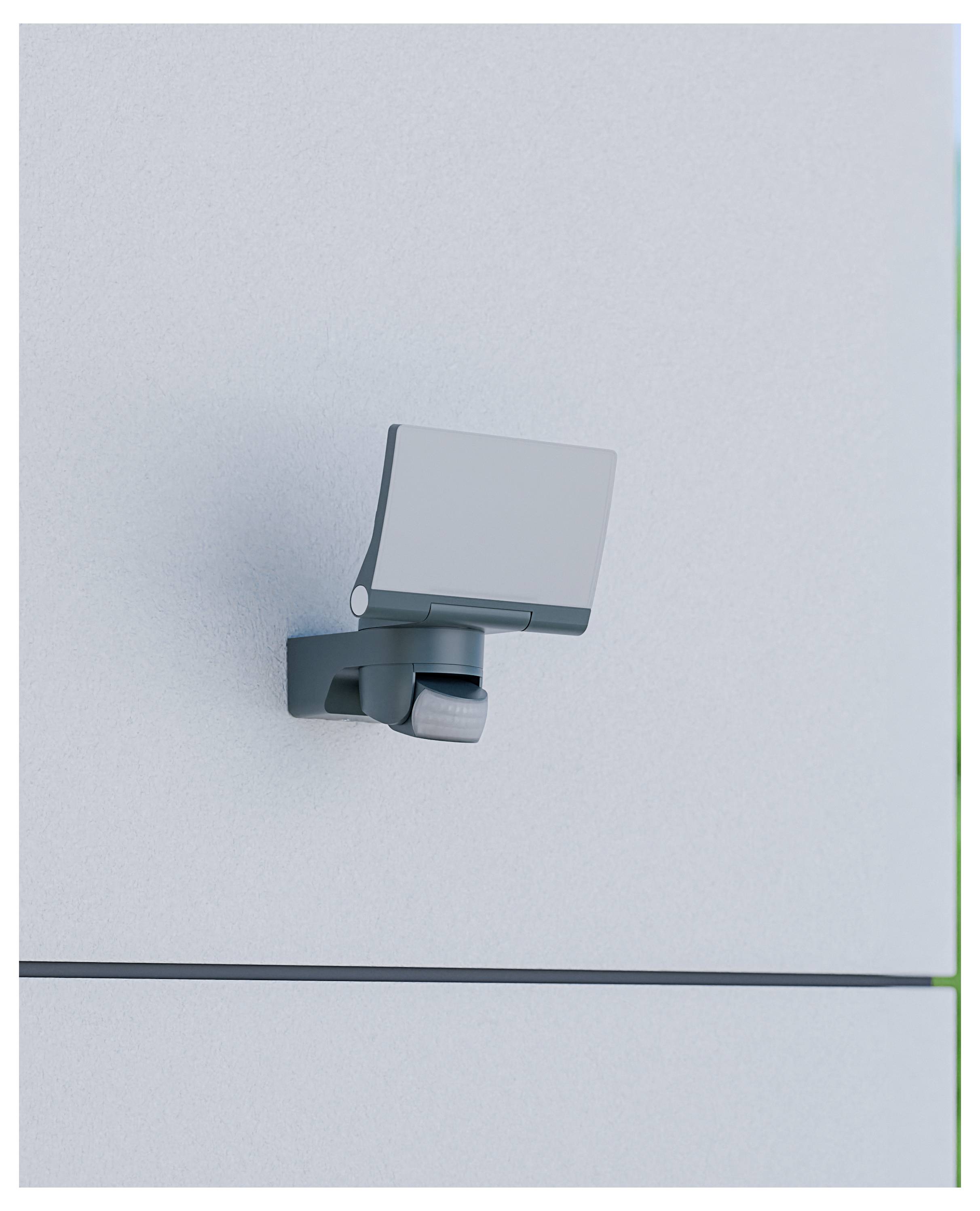 Gros plan d'une caméra murale argentée ou d'un dispositif capteur fixé sur un panneau gris clair uni.