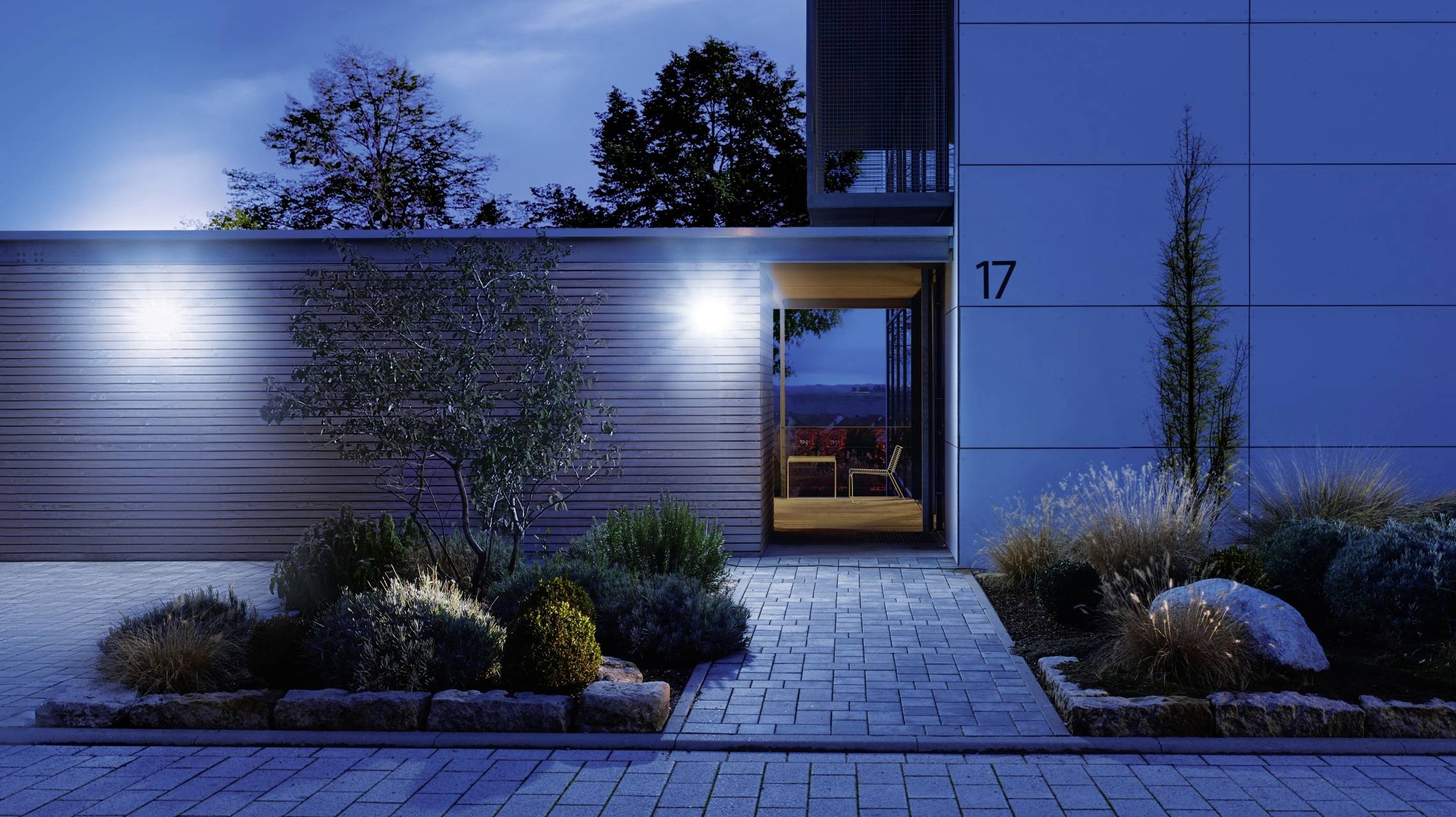 Vue extérieure moderne d'une maison la nuit, éclairée par des projecteurs. Un chemin mène à une porte en bois, entourée de plantes.
