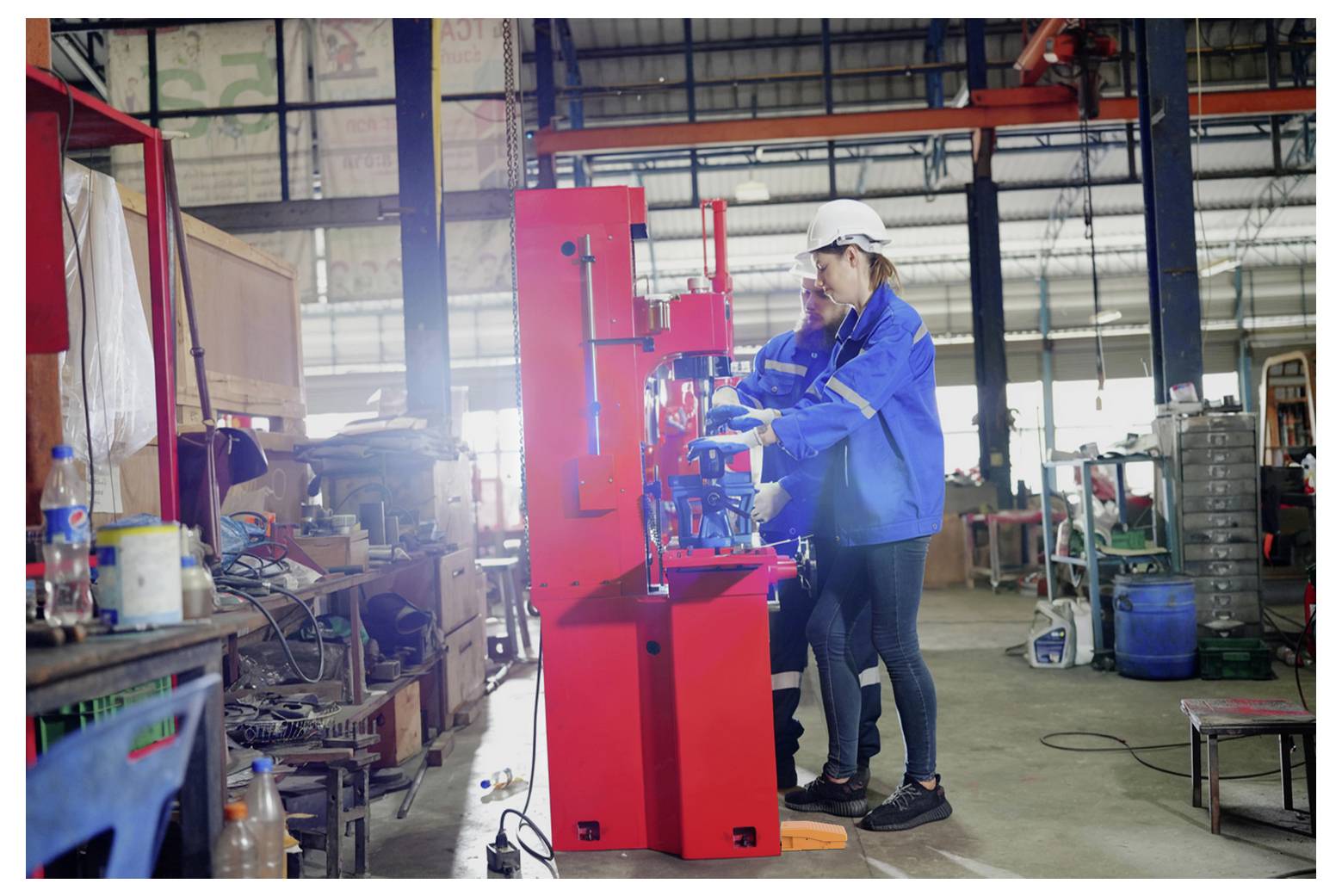 Deux ouvriers d'usine vêtus d'équipement de sécurité font fonctionner une machine industrielle rouge vif dans un atelier ou un entrepôt.