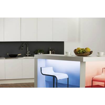 Cuisine moderne avec armoire blanche, évier et îlot central. Sur l'îlot, des tasses et un panier de fruits sont disposés. Des tabourets lumineux en bleu et rouge complètent l'ensemble.