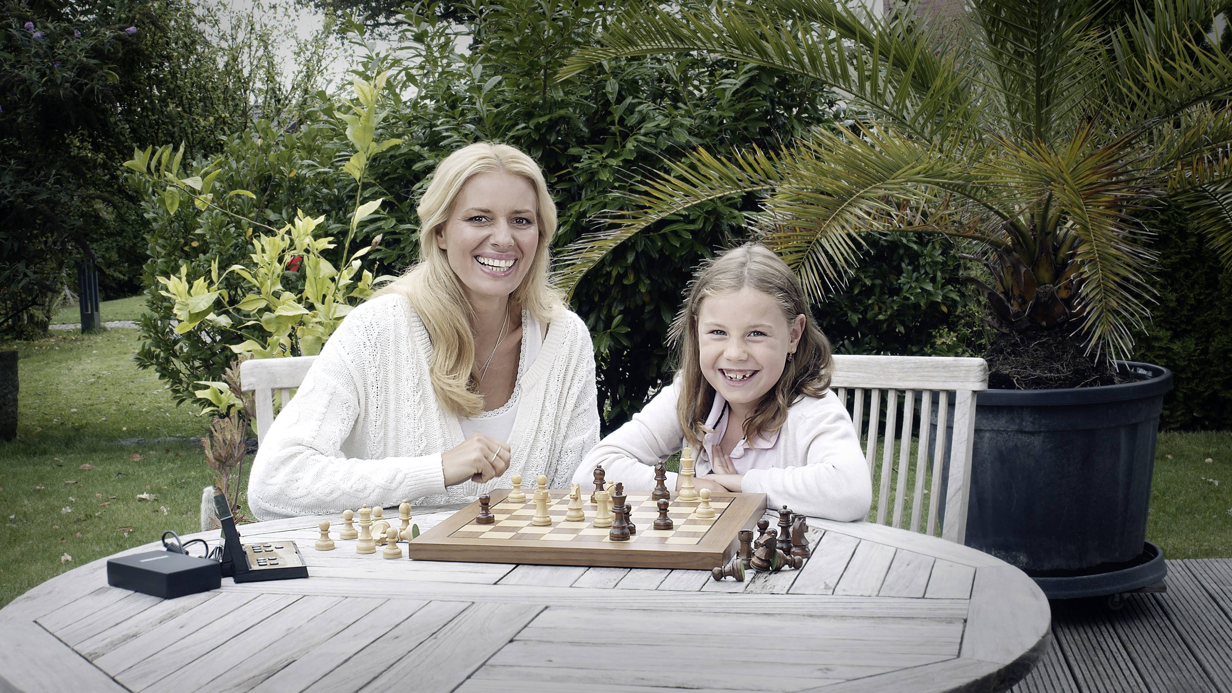 Une femme et une fille jouent aux échecs à une table ronde en bois dans le jardin, toutes deux sourient. Des plantes sont visibles en arrière-plan.