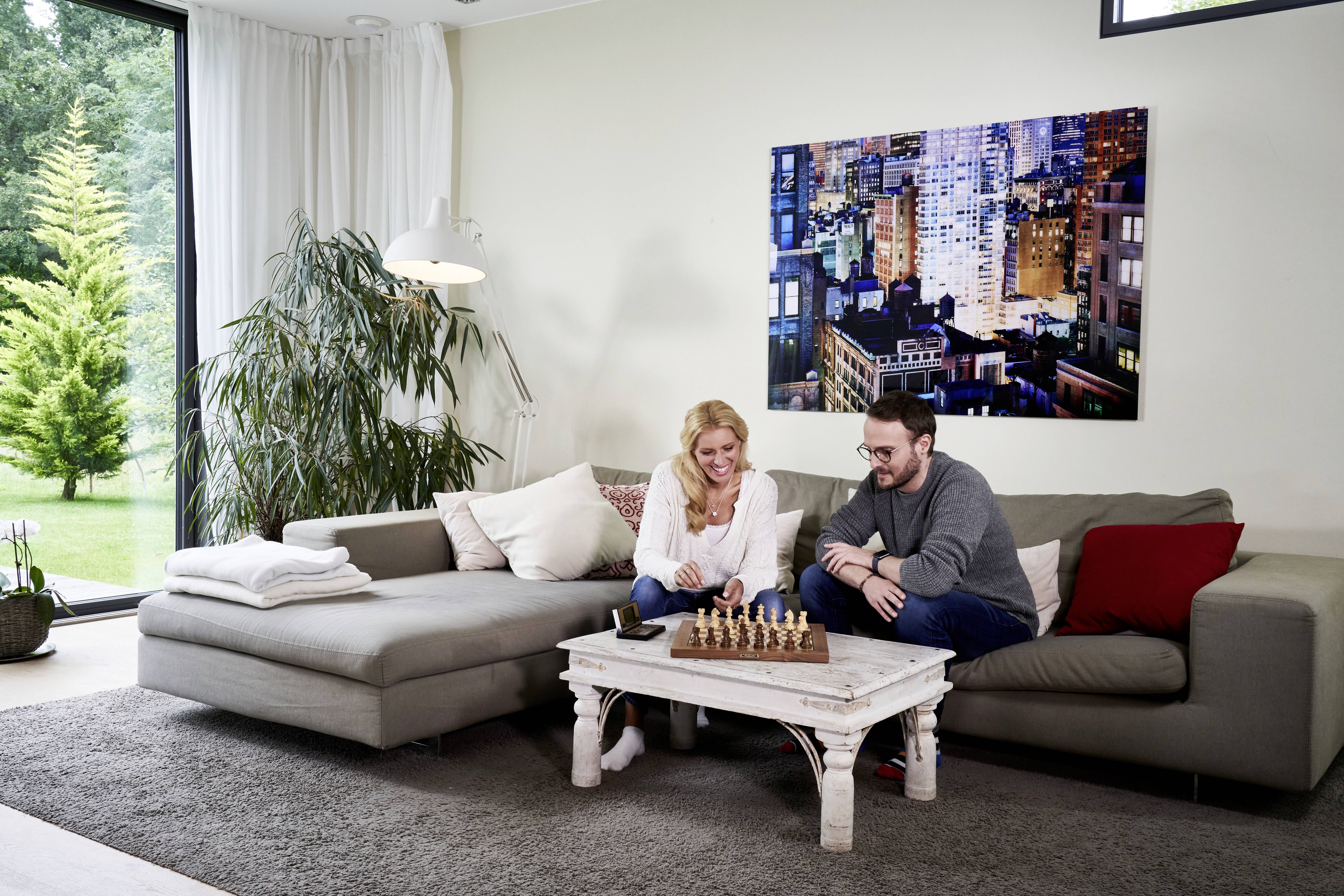 Deux personnes sont assises sur un canapé dans un salon et jouent aux échecs. En arrière-plan, on peut voir un grand paysage urbain.