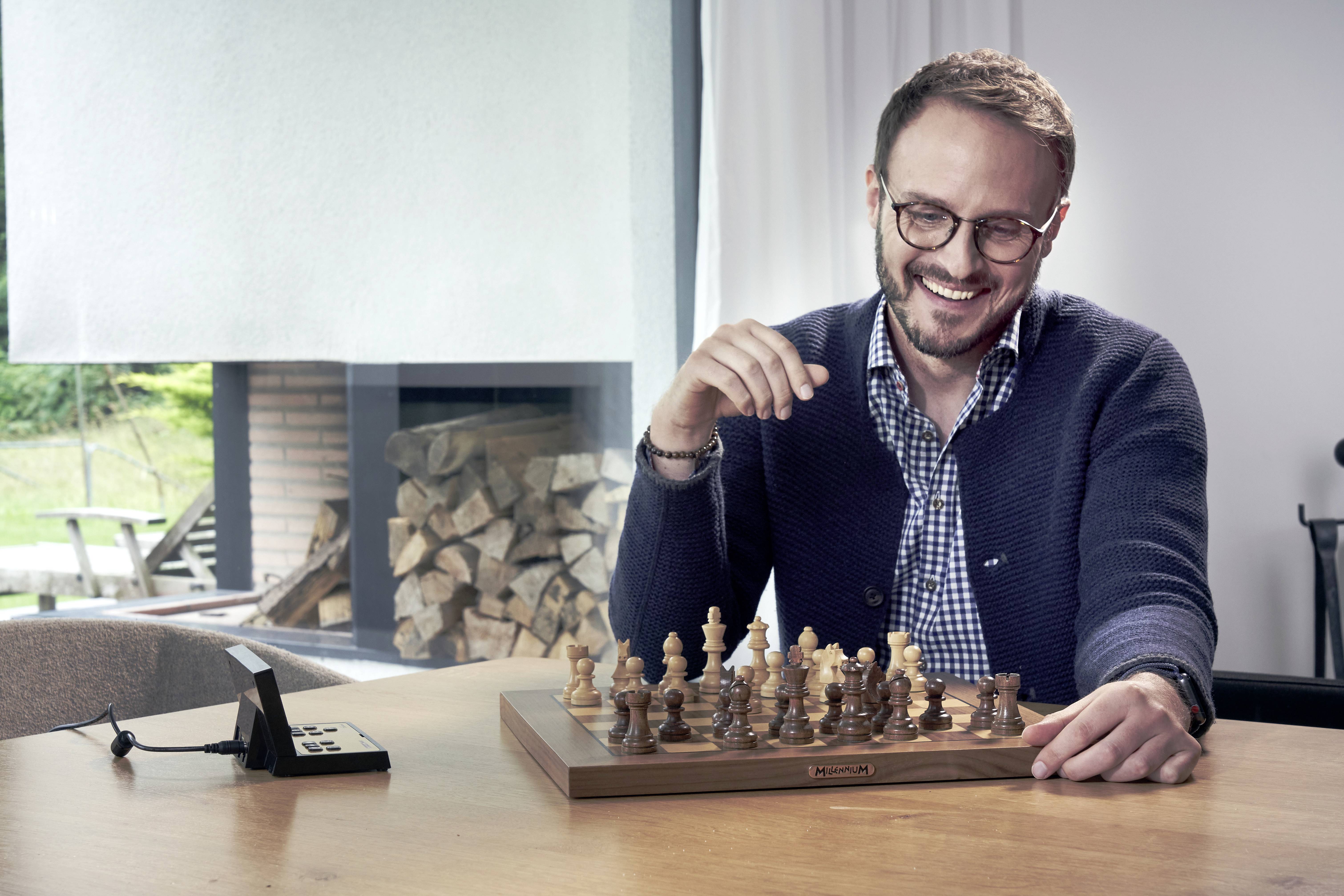 Un homme souriant est assis à la table et joue aux échecs. Derrière lui, on aperçoit une cheminée avec des bûches de bois.