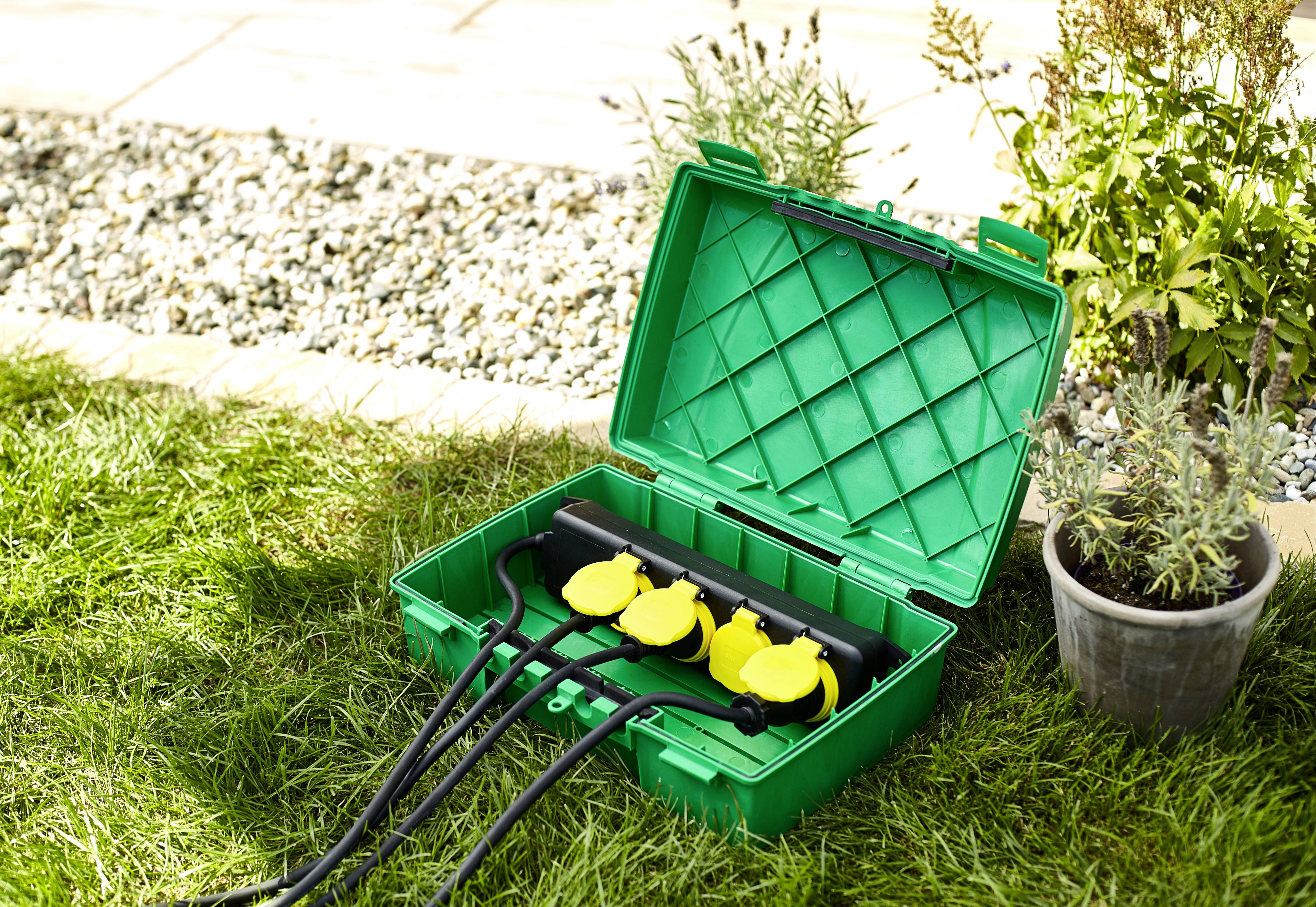 Une boîte verte ouverte avec plusieurs prises électriques jaunes et des câbles est posée sur l'herbe. À côté, on peut voir des pots de fleurs et un chemin de gravier.