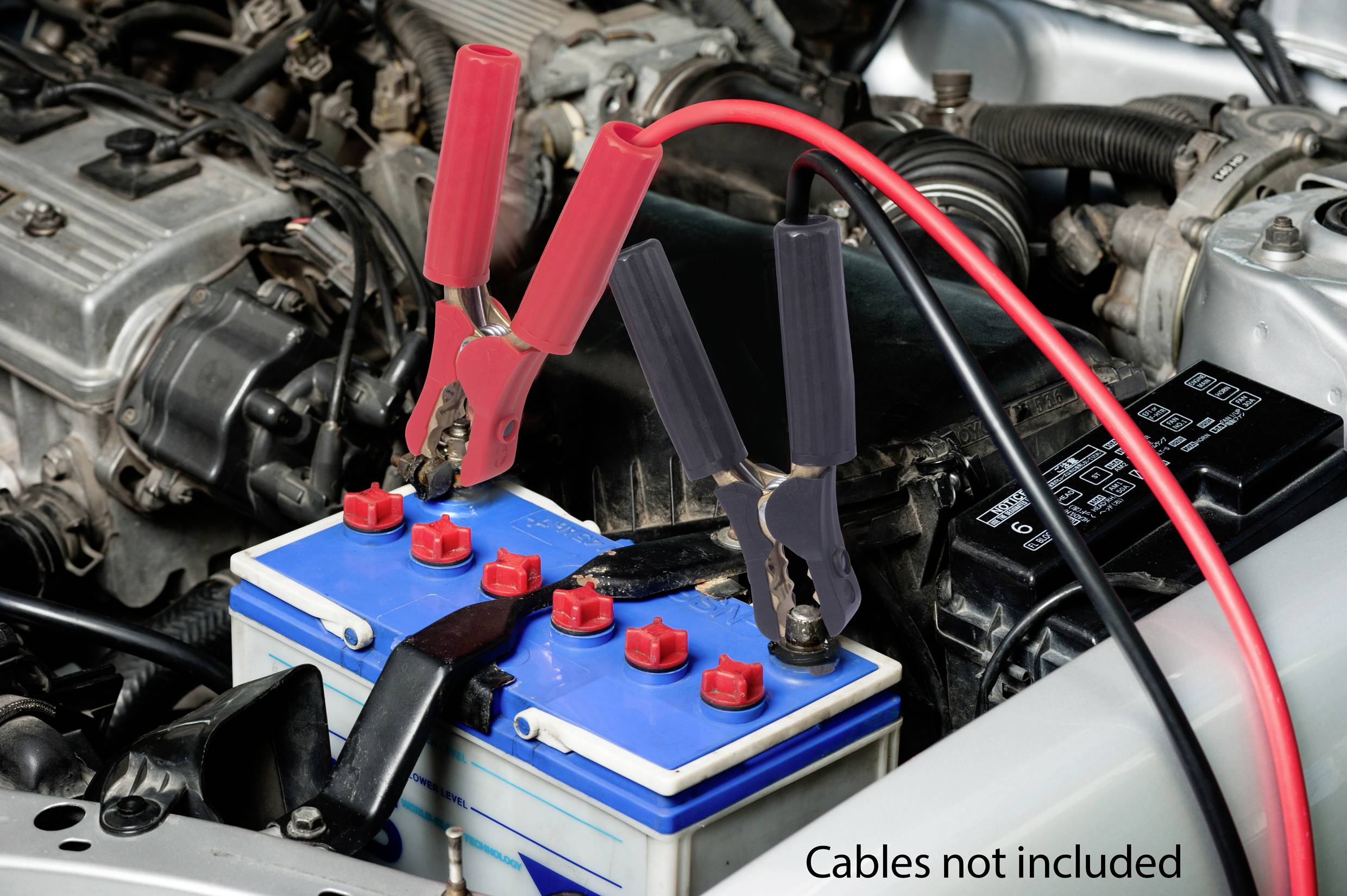 Batterie de voiture dans un compartiment moteur avec des pinces de charge connectées. Inscription sur le bord : « Câbles non fournis ».