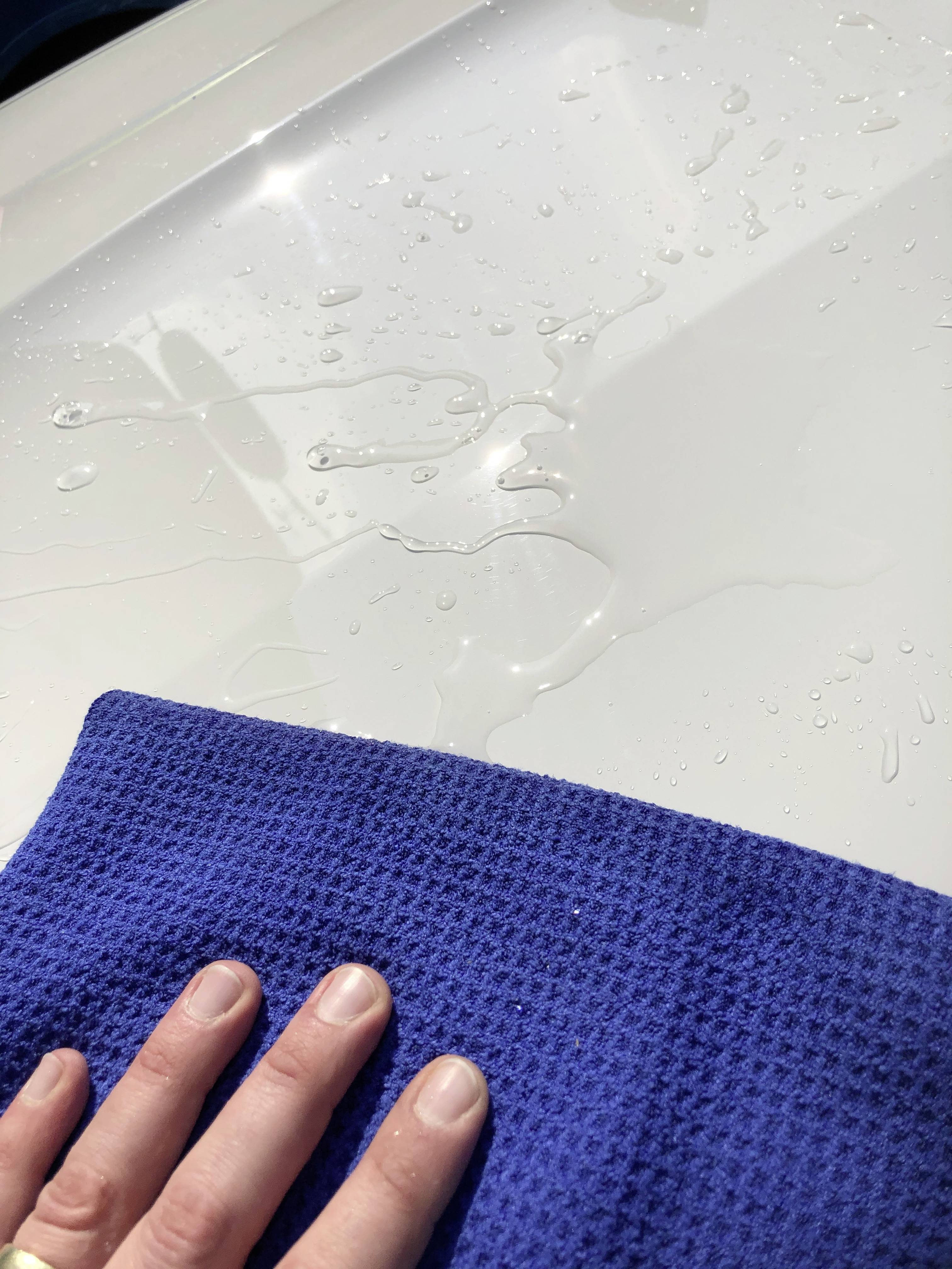 Une main avec un chiffon bleu essuie de l'eau d'une surface blanche et brillante. La lumière réfléchie indique la propreté.