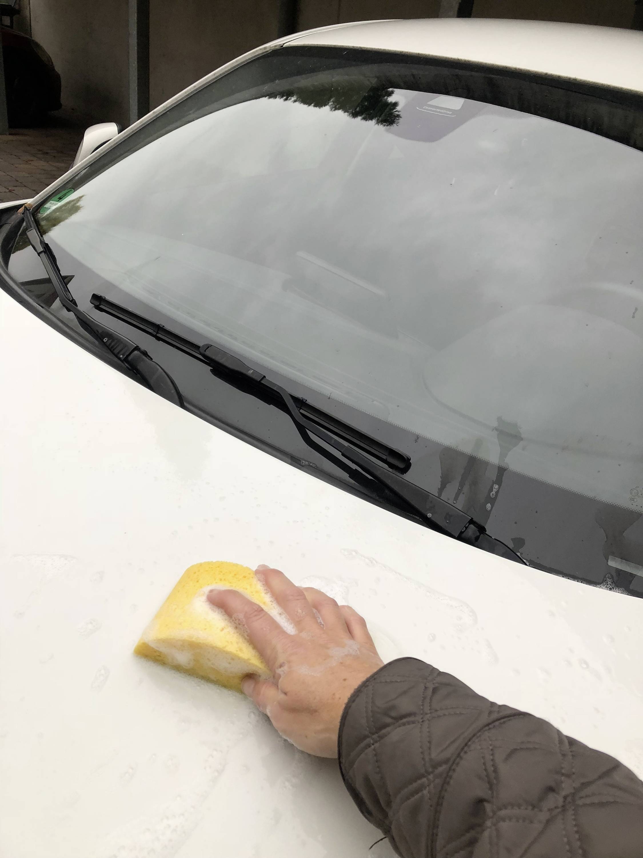 Une personne nettoie le capot d'une voiture blanche avec une éponge jaune. La voiture est garée dans une cour.