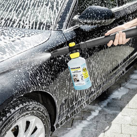 Une personne lave une voiture avec un nettoyeur haute pression relié à un réservoir de produit de nettoyage. La voiture est recouverte de mousse.