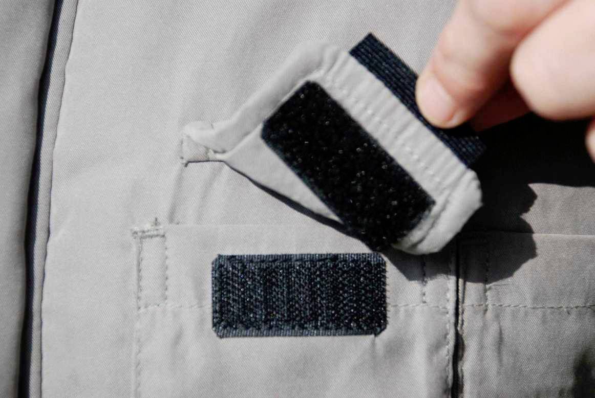 Un velcro s'ouvre. Une main soulève la partie supérieure d'un tissu gris avec des attaches auto-agrippantes.
