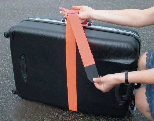 Une personne resserre une sangle orange autour d'une grande valise noire sur un sol asphalté.