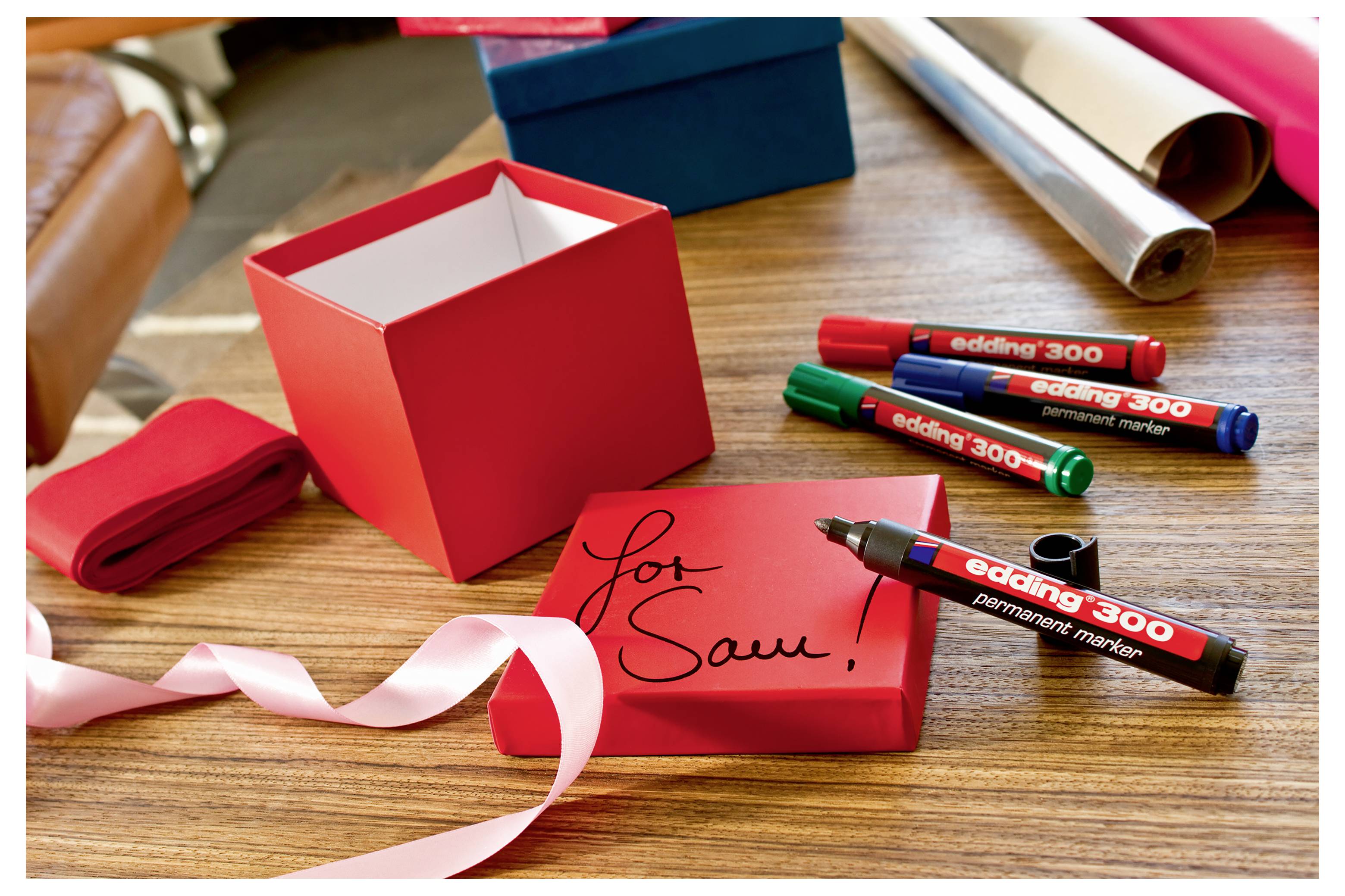 Boîte cadeau rouge avec couvercle assorti étiquetée « pour Toi ! » à côté d'un ruban et de plusieurs marqueurs de couleur sur une table en bois.