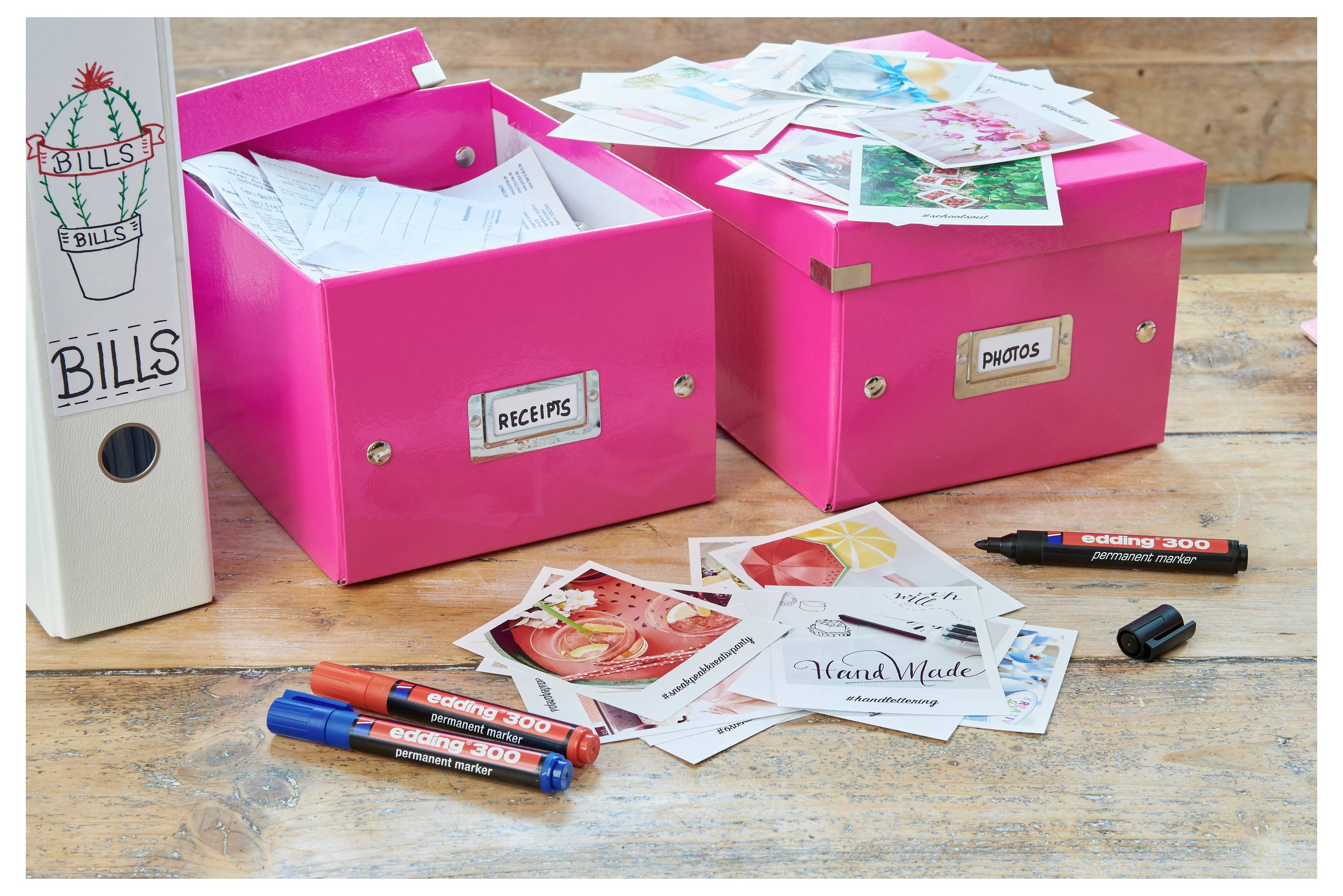Deux boîtes de rangement rose vif étiquetées « Reçus » et « Photos » sur une table en bois, avec des reçus imprimés éparpillés et des cartes photo, plus un classeur blanc « Factures » et des stylos.