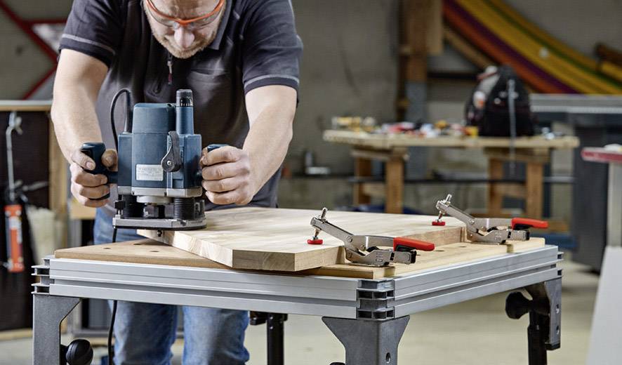Une personne usine une planche de bois à l'aide d'une fraiseuse dans un atelier. Des serre-joints de table maintiennent le bois en place. Des outils sont visibles en arrière-plan.