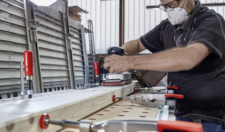 Un homme ponce une planche en bois avec une ponceuse dans un atelier. Il porte des lunettes de protection et un masque.