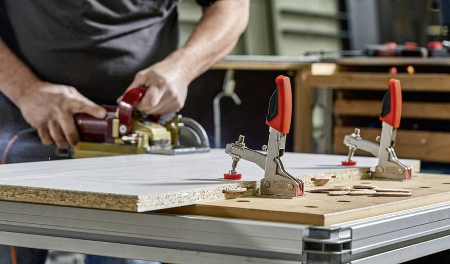Une personne ponce une planche de bois fixée sur une table de travail à l'aide de deux serre-joints. Des outils sont visibles en arrière-plan.