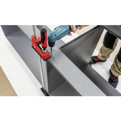 Une personne monte un meuble avec une perceuse. Un élément de structure est maintenu à l'aide d'un serre-joint rouge.