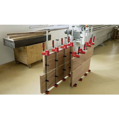 Des planches de bois sont maintenues ensemble par plusieurs pinces rouges dans un atelier. À l'arrière-plan, on peut voir des machines et des outils.