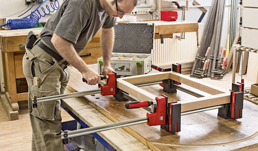Un homme travaille dans un atelier et utilise des serre-joints pour maintenir des pièces de cadre sur une table. Des outils et des matériaux sont disposés autour de lui.