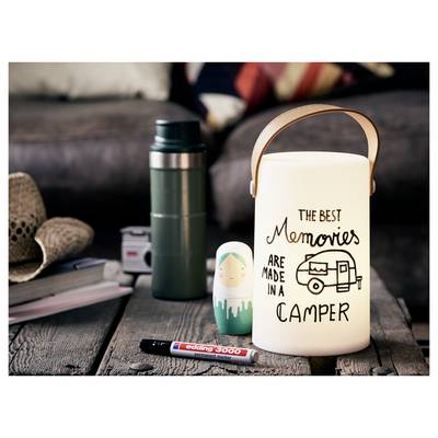 Un porte-bougie de style lanterne affichant le texte « Les meilleurs souvenirs se font en camping-car » est assis avec un thermos, un marqueur et une poupée russe sur une table rustique.