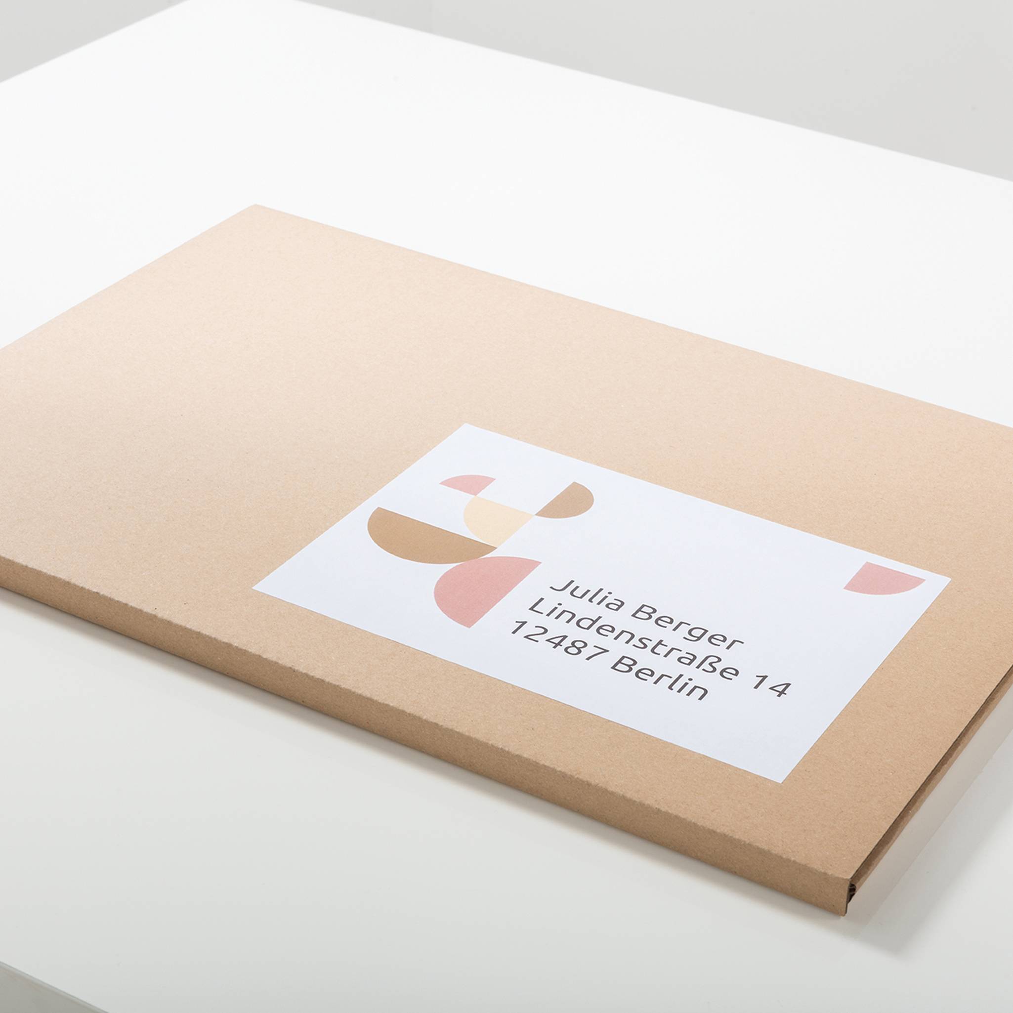 Une enveloppe marron rectangulaire et plate, portant une étiquette avec des formes colorées et l'adresse « Julia Berger, Lindenstraße 14, 12487 Berlin ».