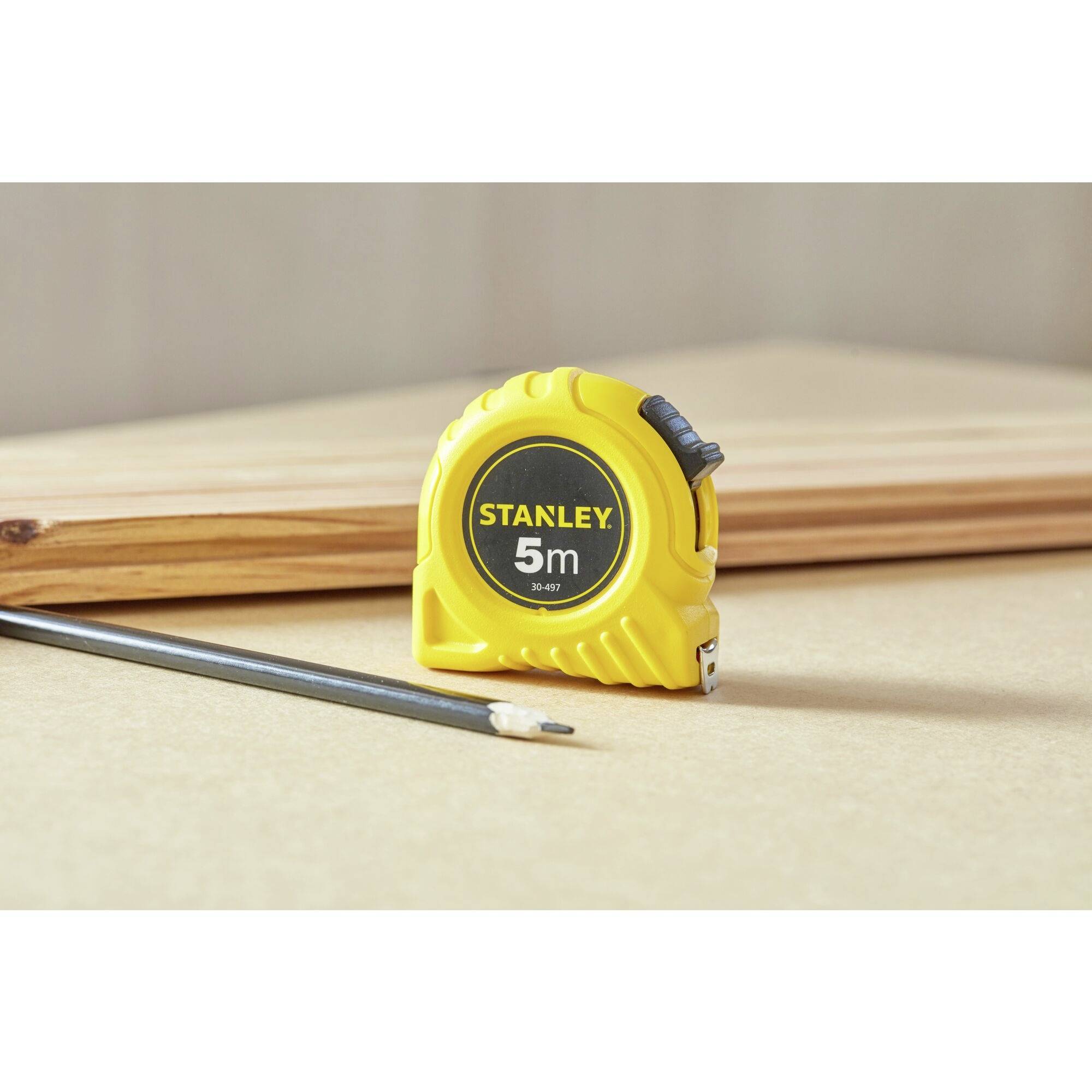 Un mètre ruban avec un boîtier jaune est posé sur une table à côté d'un crayon et d'un morceau de bois. Sur le mètre ruban, il est inscrit 'Stanley 5m'.