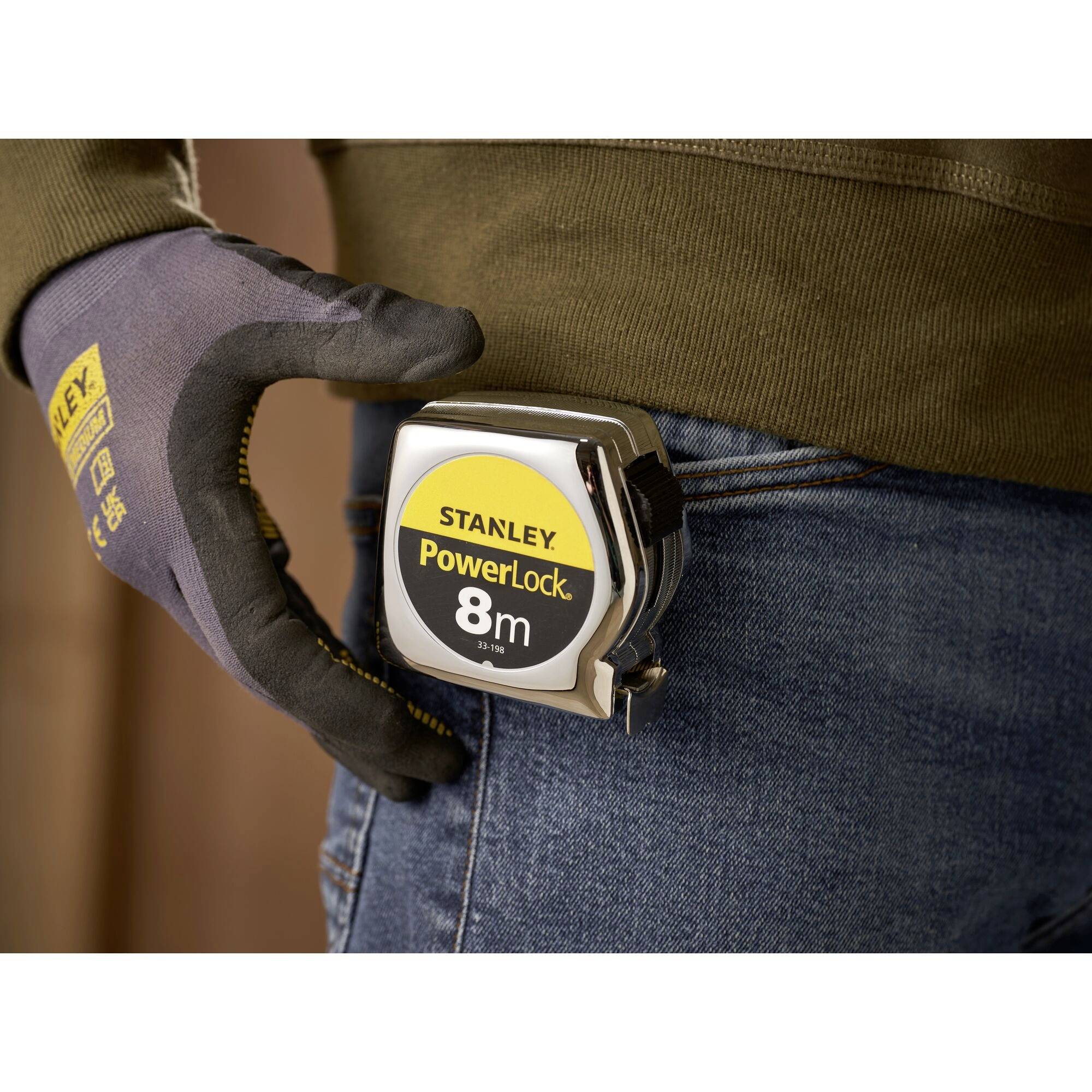 Un gant tient un mètre ruban portant l'inscription 'Stanley PowerLock 8m', attaché à la ceinture d'une personne.