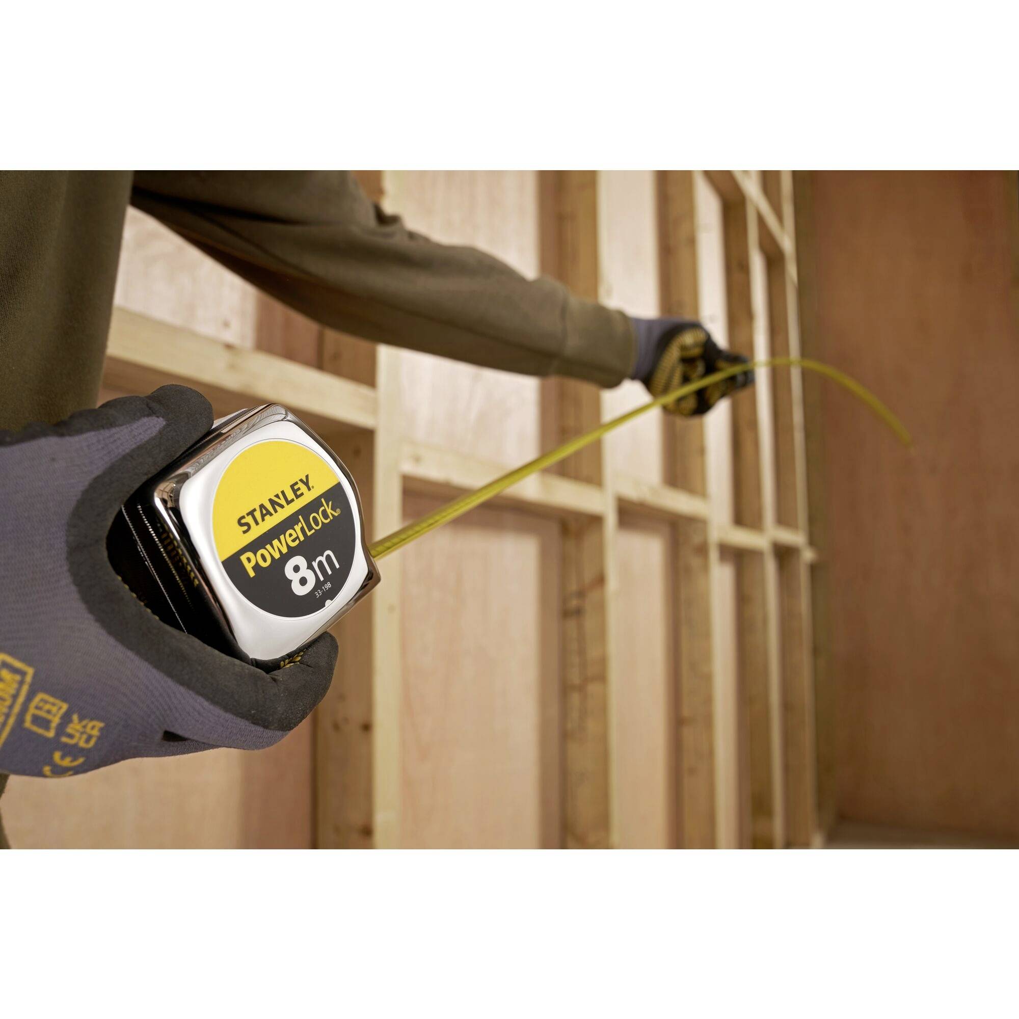 Une personne portant des gants mesure une construction en bois à l'aide d'un mètre ruban de 8 mètres.