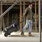 Un homme tire une valise à outils noire sur roulettes à travers un chantier avec des structures en bois. Il porte un jean et des chaussures de travail.
