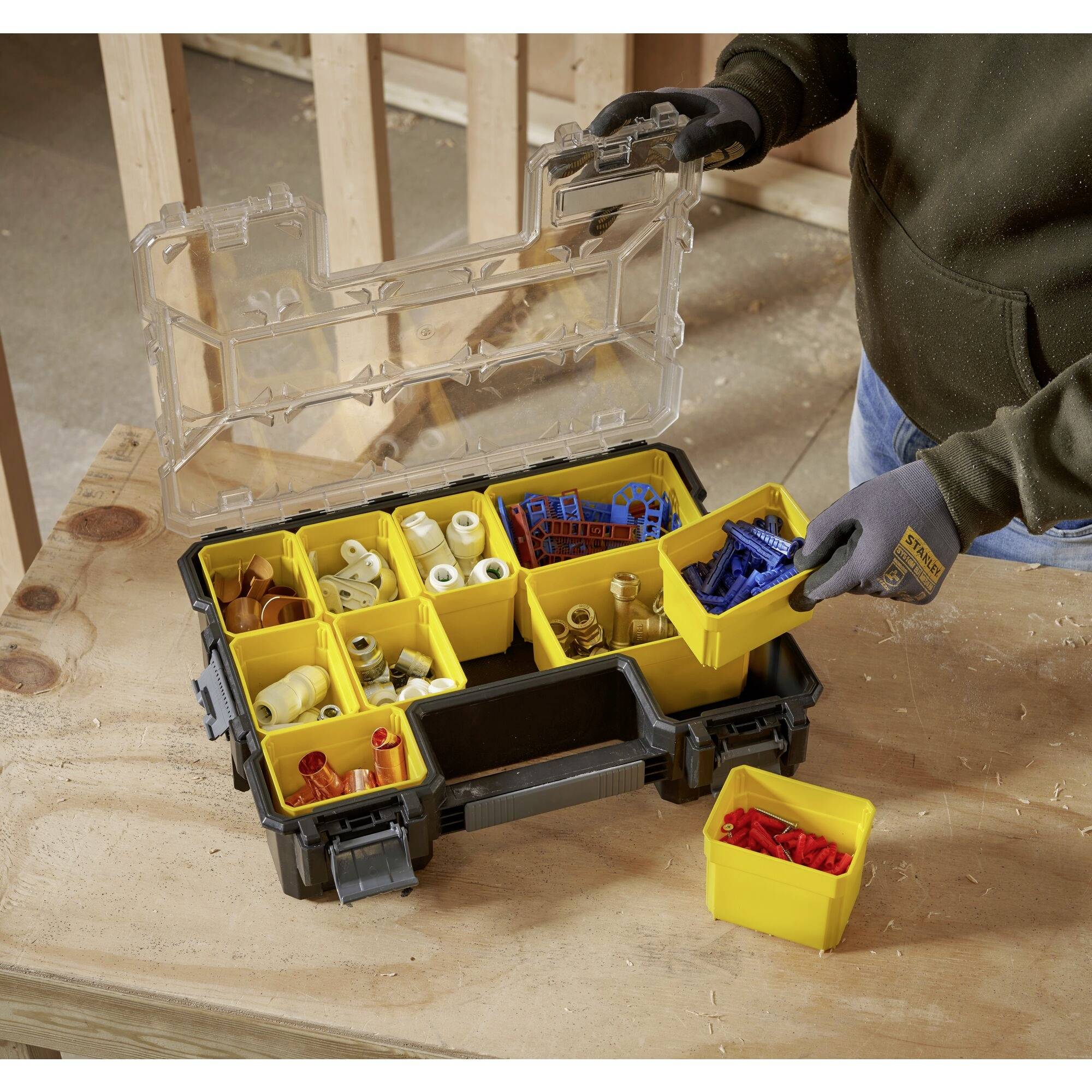 Une boîte à outils ouverte avec des compartiments jaunes, remplie de divers éléments de fixation, est posée sur une table en bois. Une personne en retire un récipient.