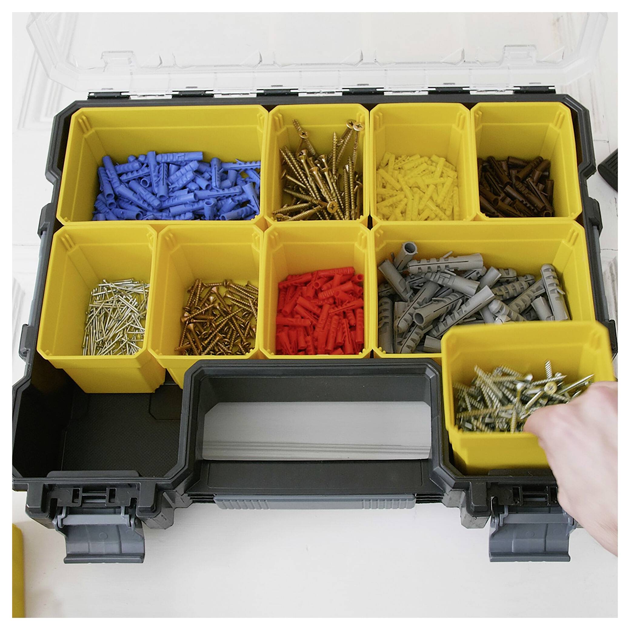 Une boîte à outils ouverte avec plusieurs compartiments. Contient différentes vis et chevilles de couleurs et de tailles variées. Une main se saisit d'un compartiment jaune.