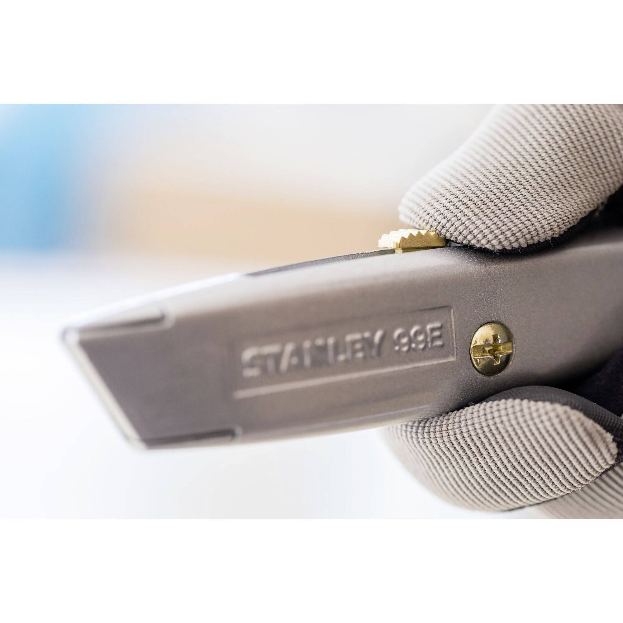 Une main gantée de gris tient un couteau argenté portant l'inscription 'STANLEY 99E'. Lame tranchante visible, arrière-plan flou.