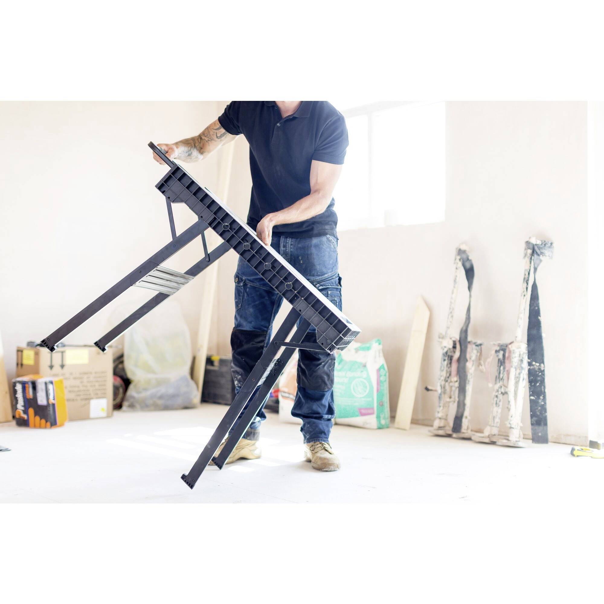 Un homme tient une table pliante dans une pièce lumineuse. Des outils et des matériaux de construction sont visibles en arrière-plan.