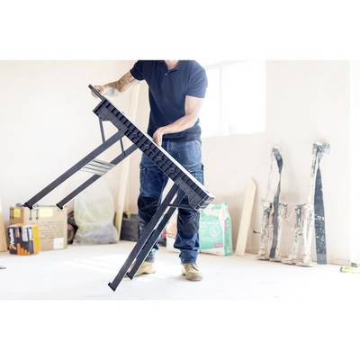 Un homme tient une table pliante dans une pièce lumineuse. Des outils et des matériaux de construction sont visibles en arrière-plan.