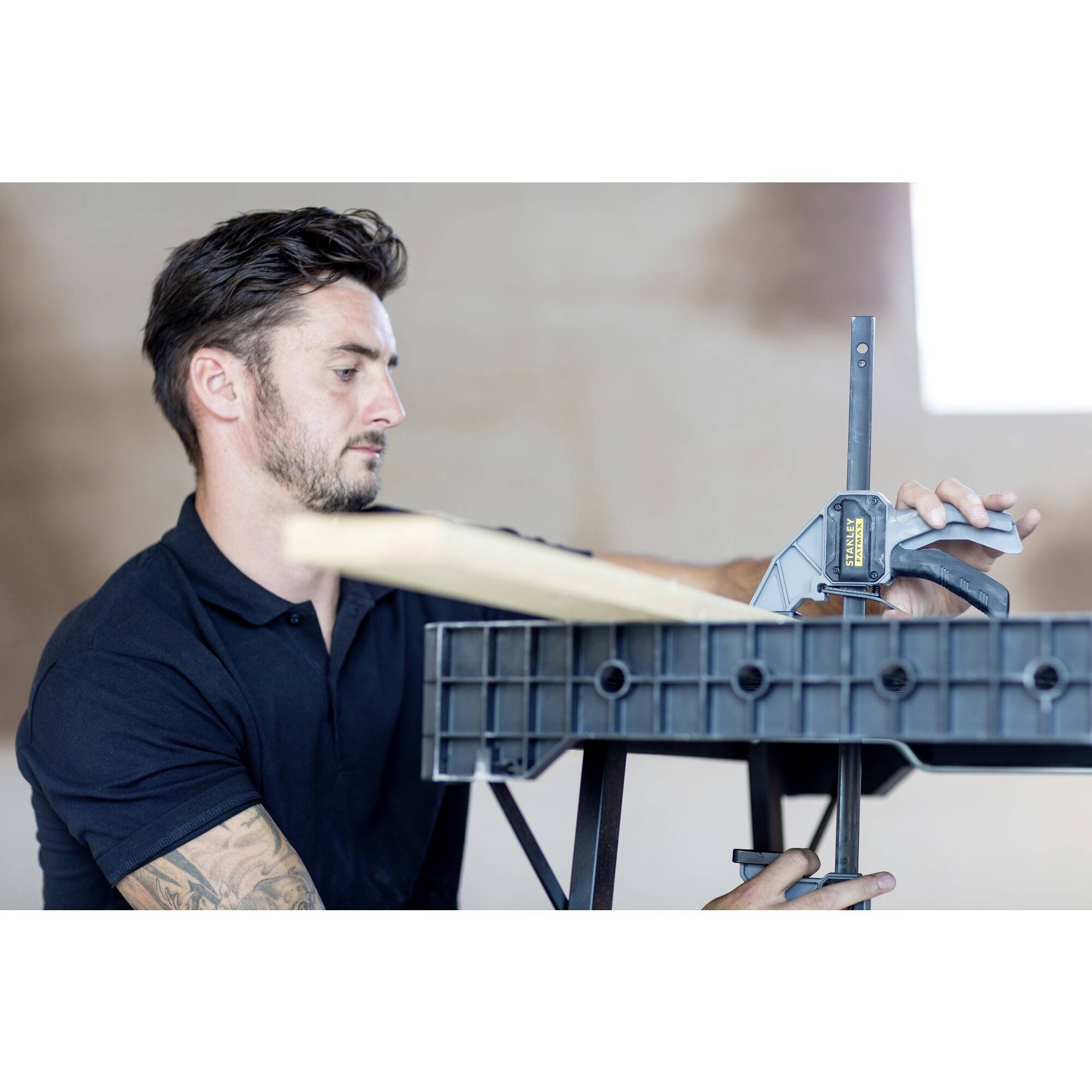 Un homme portant un t-shirt noir travaille avec concentration à un établi, utilisant une pince pour maintenir un morceau de bois.