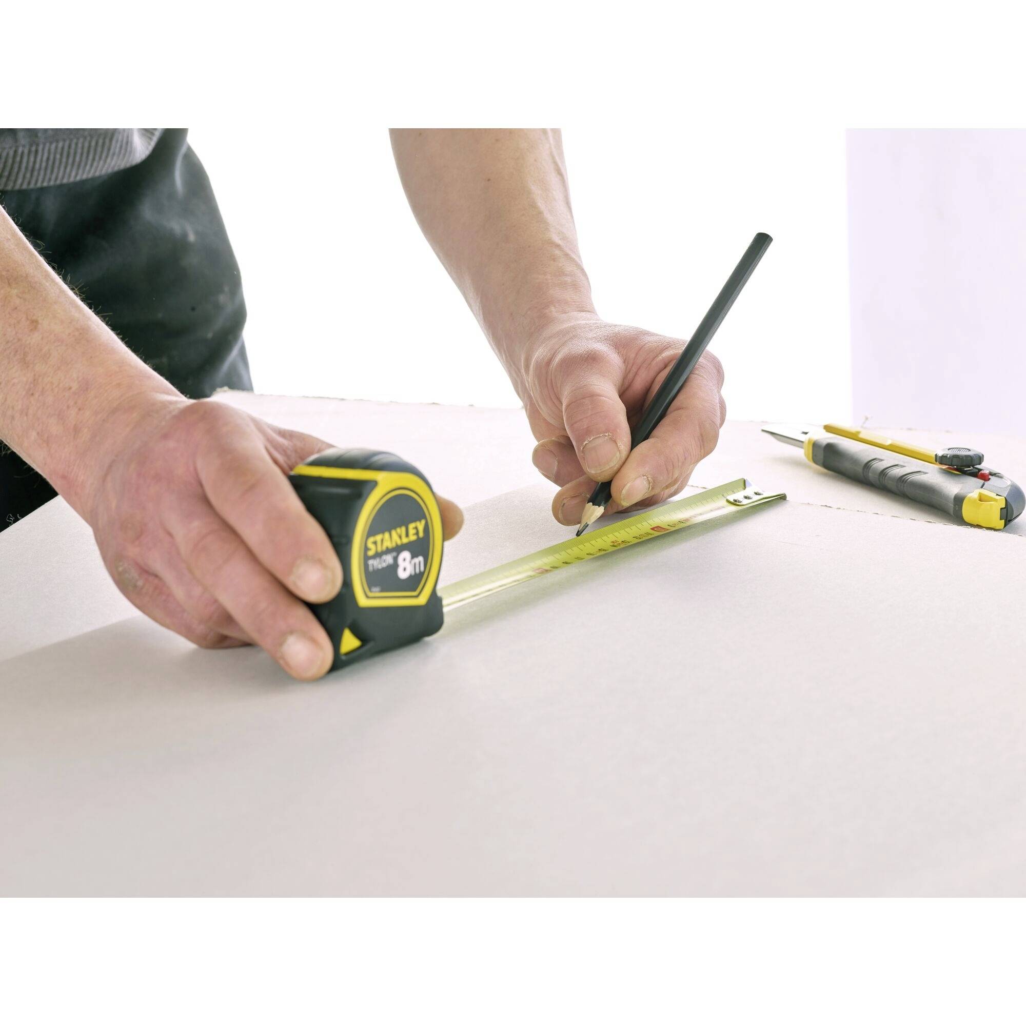 Une personne mesure une surface en bois à l'aide d'un mètre ruban de 8 mètres et marque un point avec un crayon. Des outils sont posés à côté.