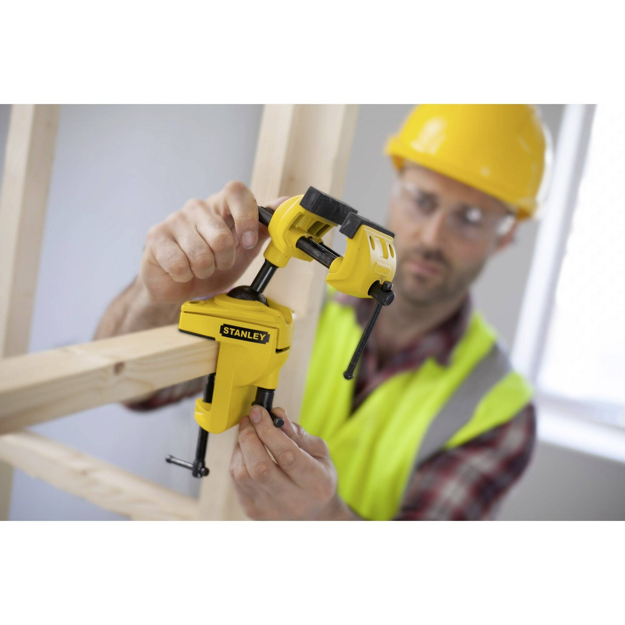 Un ouvrier portant un casque jaune et un gilet de sécurité fixe une pince jaune sur une planche de bois dans une pièce en construction.