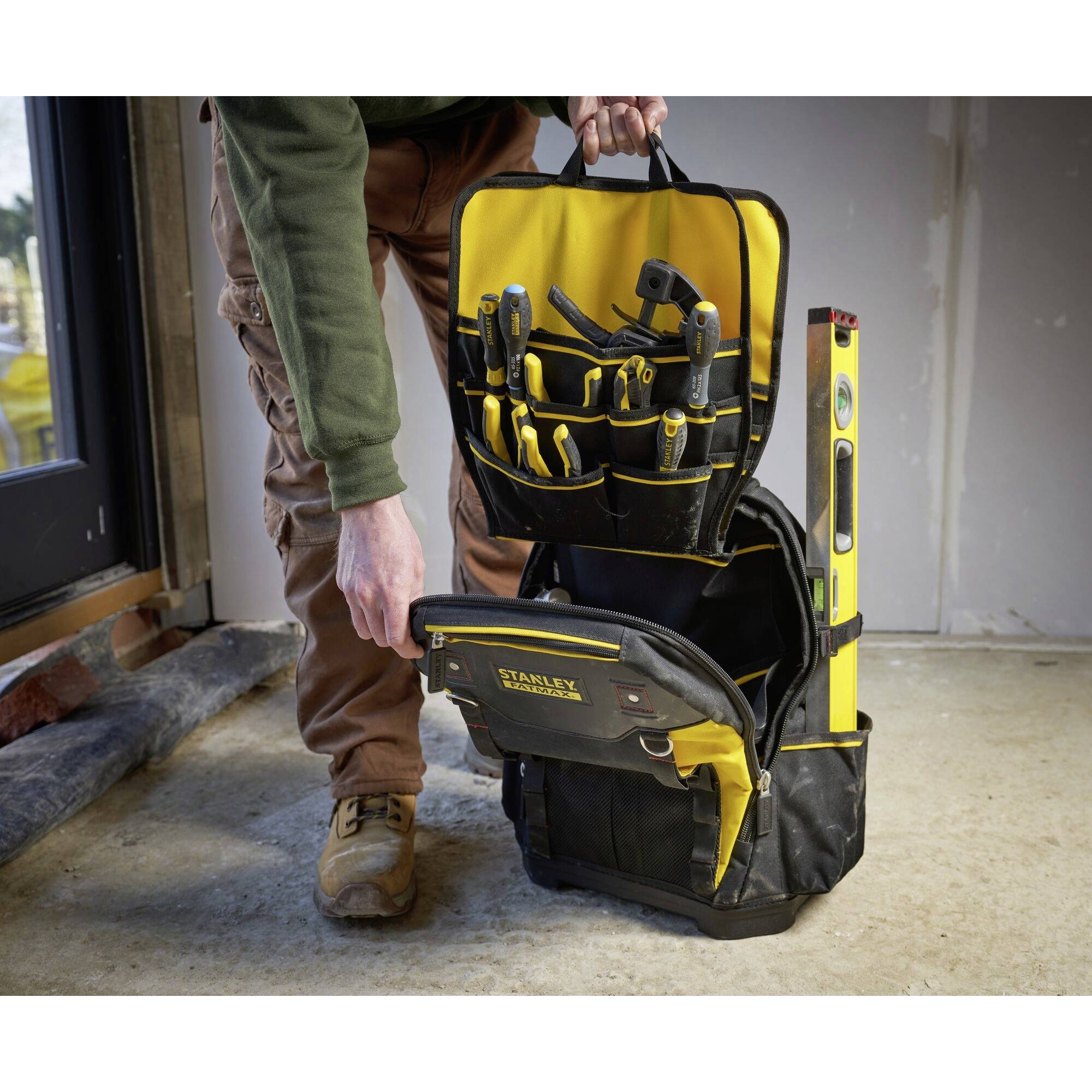 Une personne tient un sac à outils rempli d'instruments. Un niveau à bulle dépasse du sac. La scène se déroule dans une pièce inachevée.