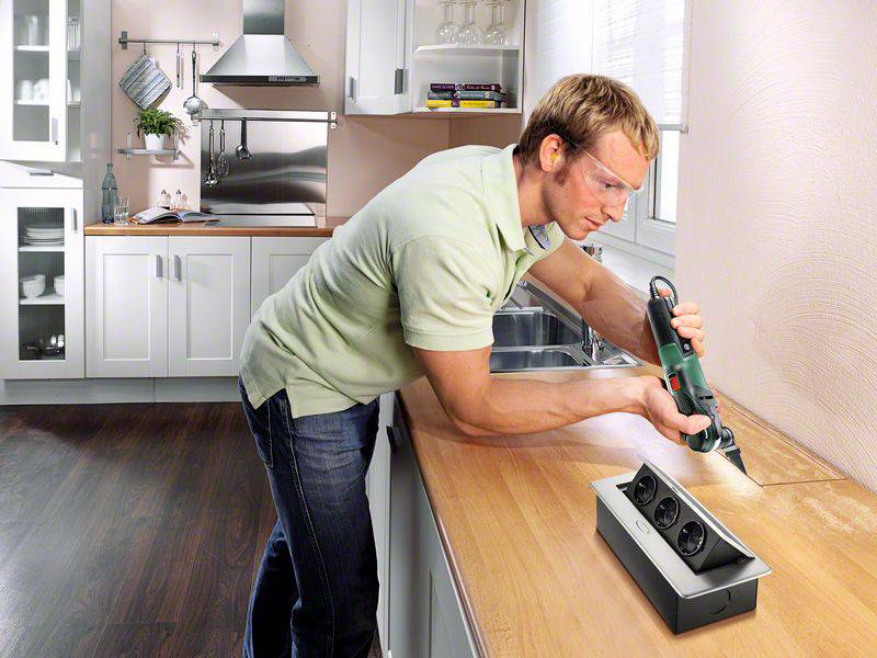 Un homme travaille avec un outil électrique sur un plan de travail de cuisine. Il porte des lunettes de protection et se trouve dans une cuisine moderne.