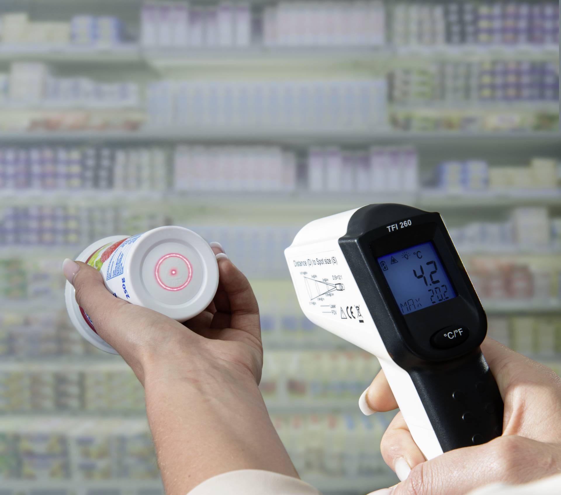 Une personne mesure la température d'un pot de yaourt à l'aide d'un thermomètre infrarouge devant un rayon réfrigéré dans un supermarché.
