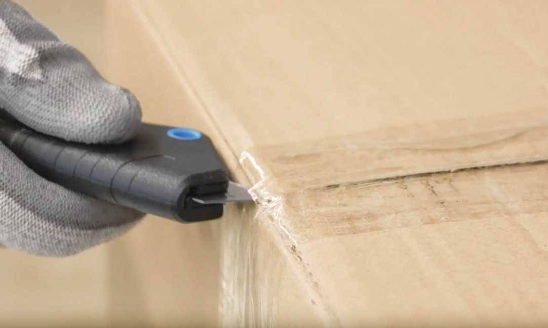 Une main gantée tient un couteau de sécurité noir et ouvre une boîte en carton.