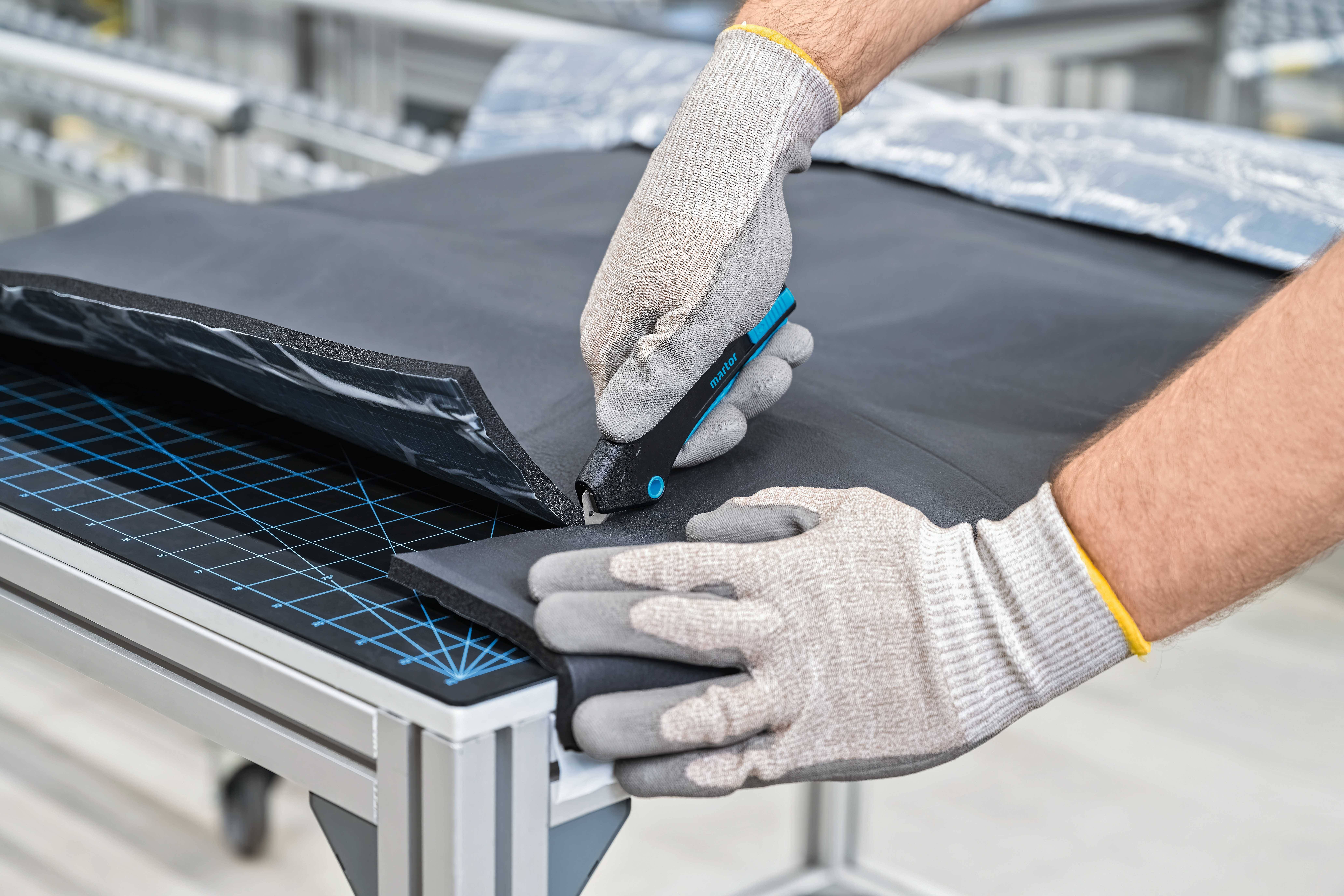 Une personne coupe un matériau noir avec un cutter sur une table de découpe, portant des gants de travail.