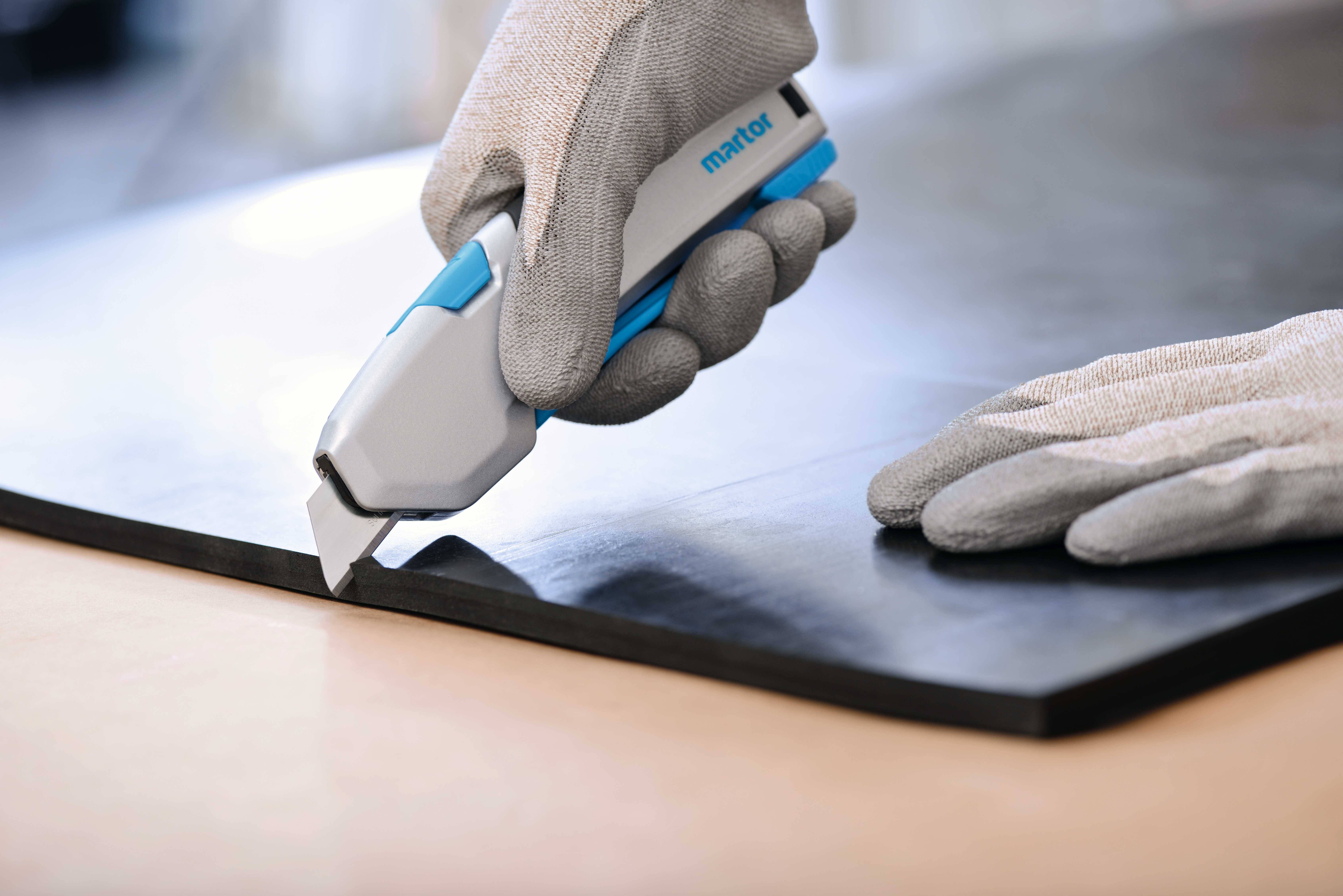 Une personne coupe un tapis noir à l'aide d'un couteau de sécurité. Les deux mains sont gantées de gants de travail gris.
